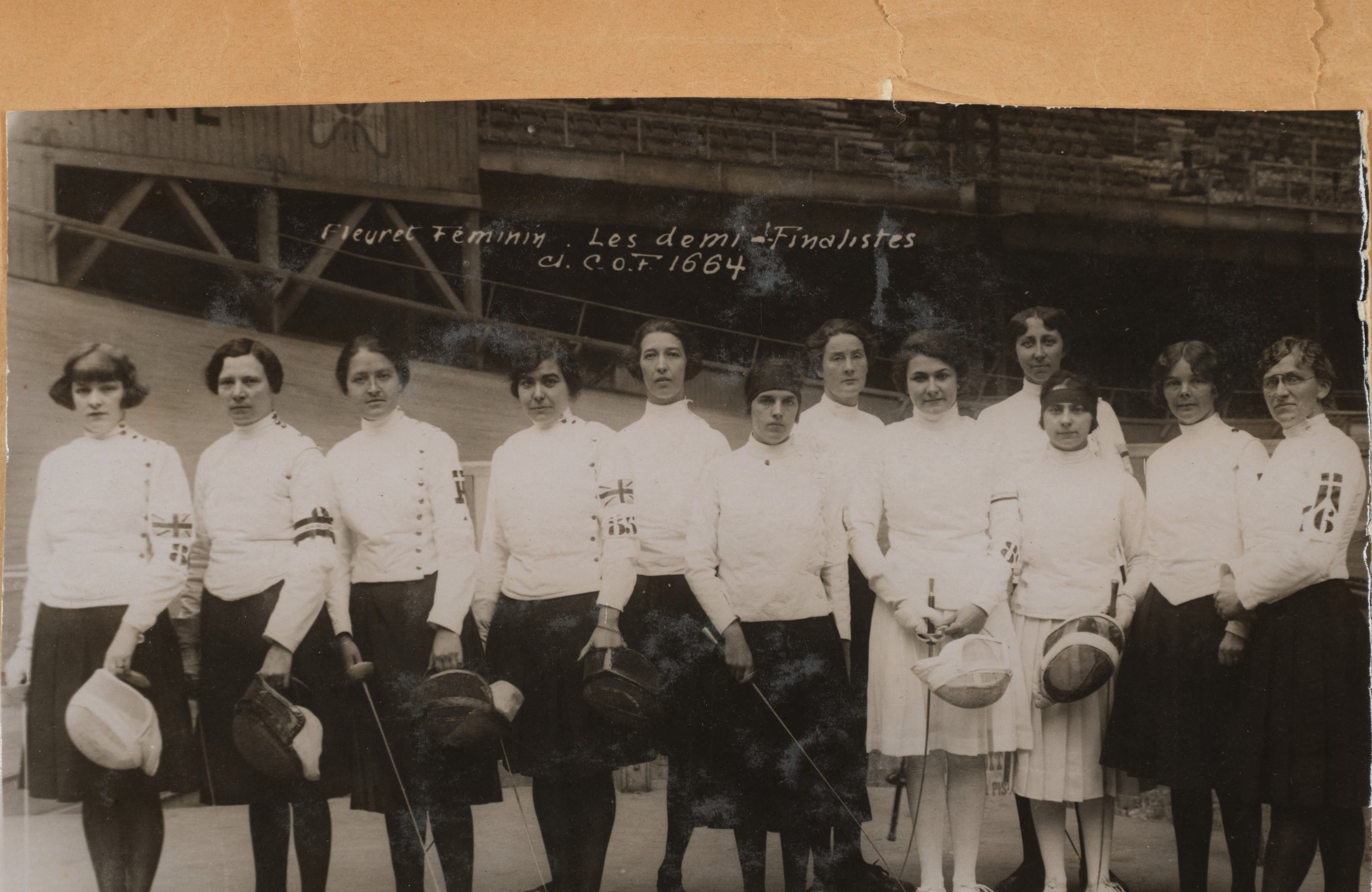 A women's historical sports team poses for a group photo, each wearing fencing attire and holding their masks. They stand in front of a wooden structure