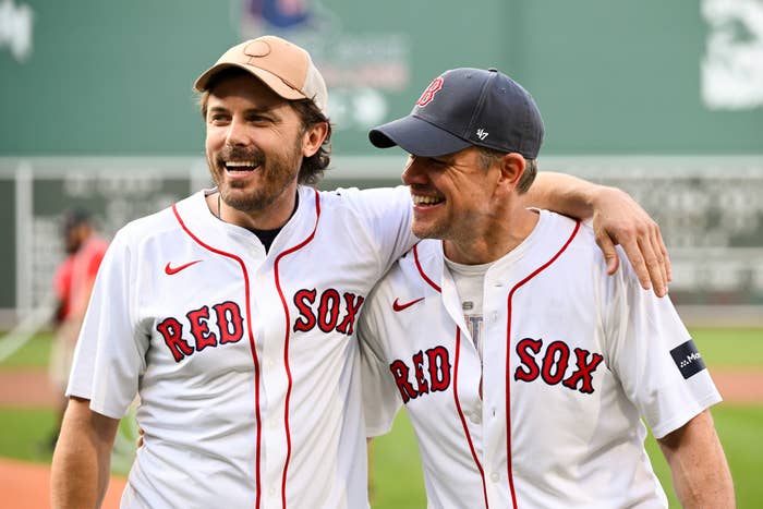 Casey Affleck and Matt Damon, wearing Red Sox jerseys and baseball caps, smiling with arms around each other's shoulders on a baseball field