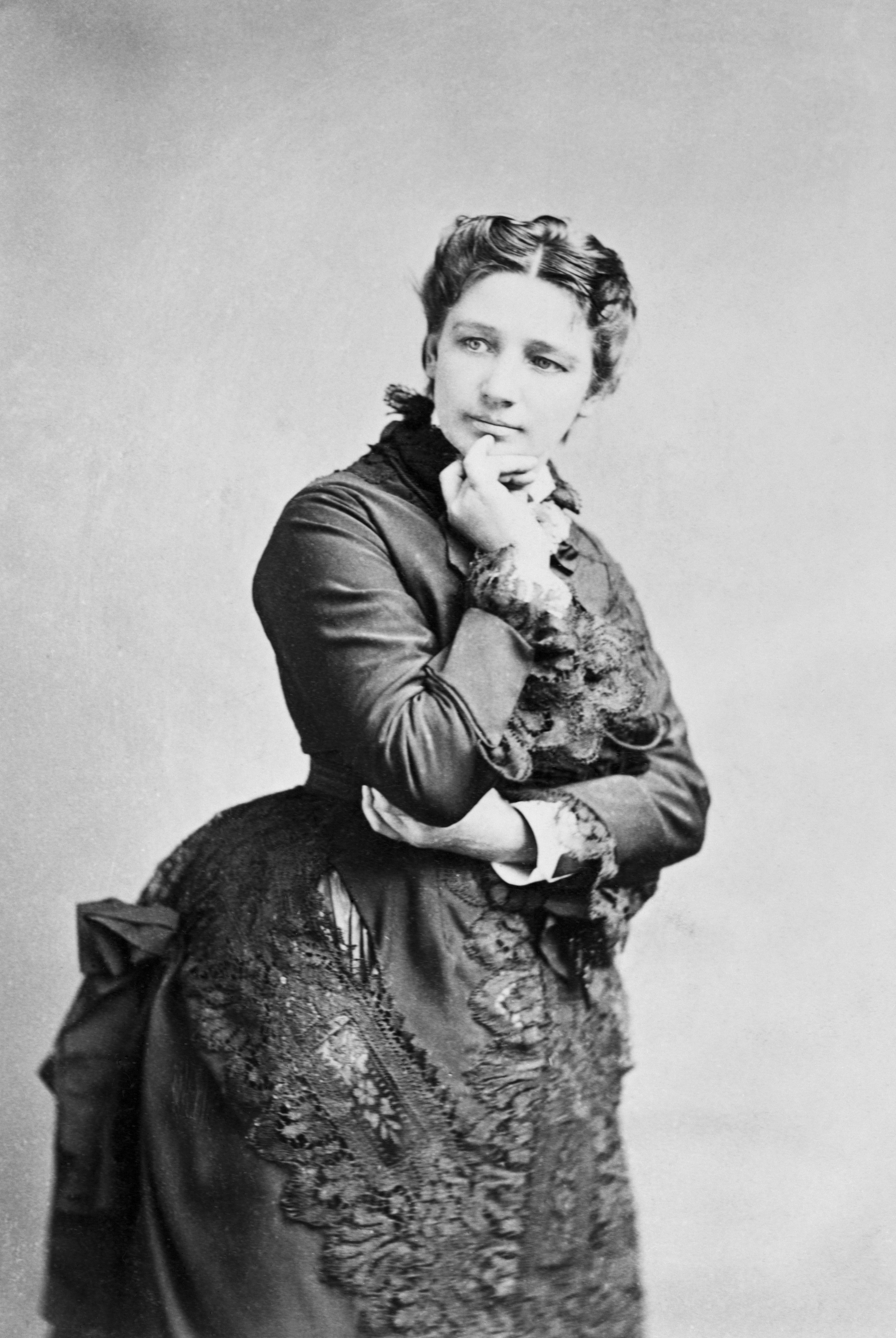 A historical portrait of Victoria Woodhull, wearing a Victorian-era dress with lace details, with her hand resting on her chin in a thoughtful pose