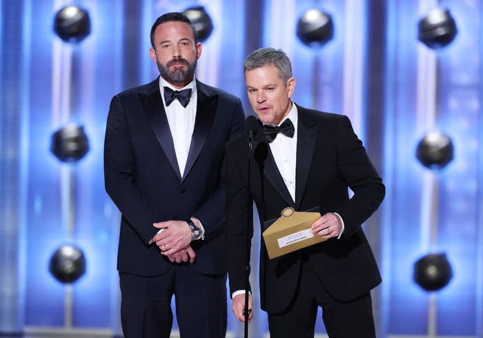 Ben Affleck and Matt Damon on stage in tuxedos, speaking at an event while Matt holds an envelope
