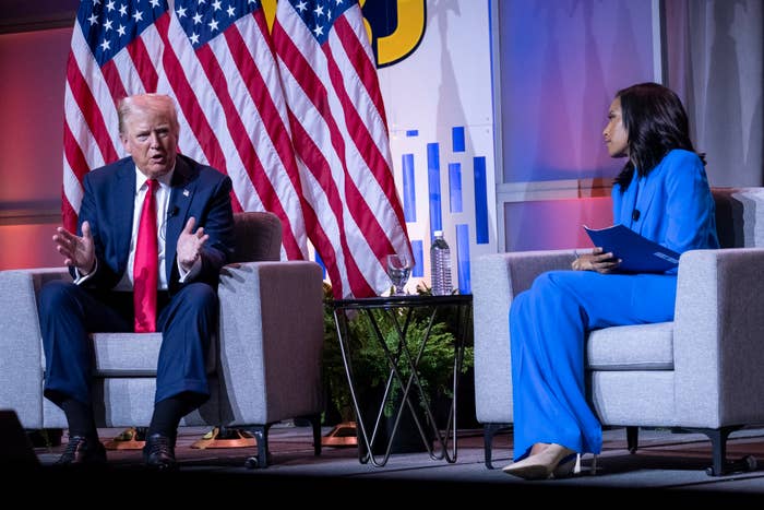 Donald Trump speaks while seated on stage next to a woman in a blue suit holding a folder. Two American flags are in the background