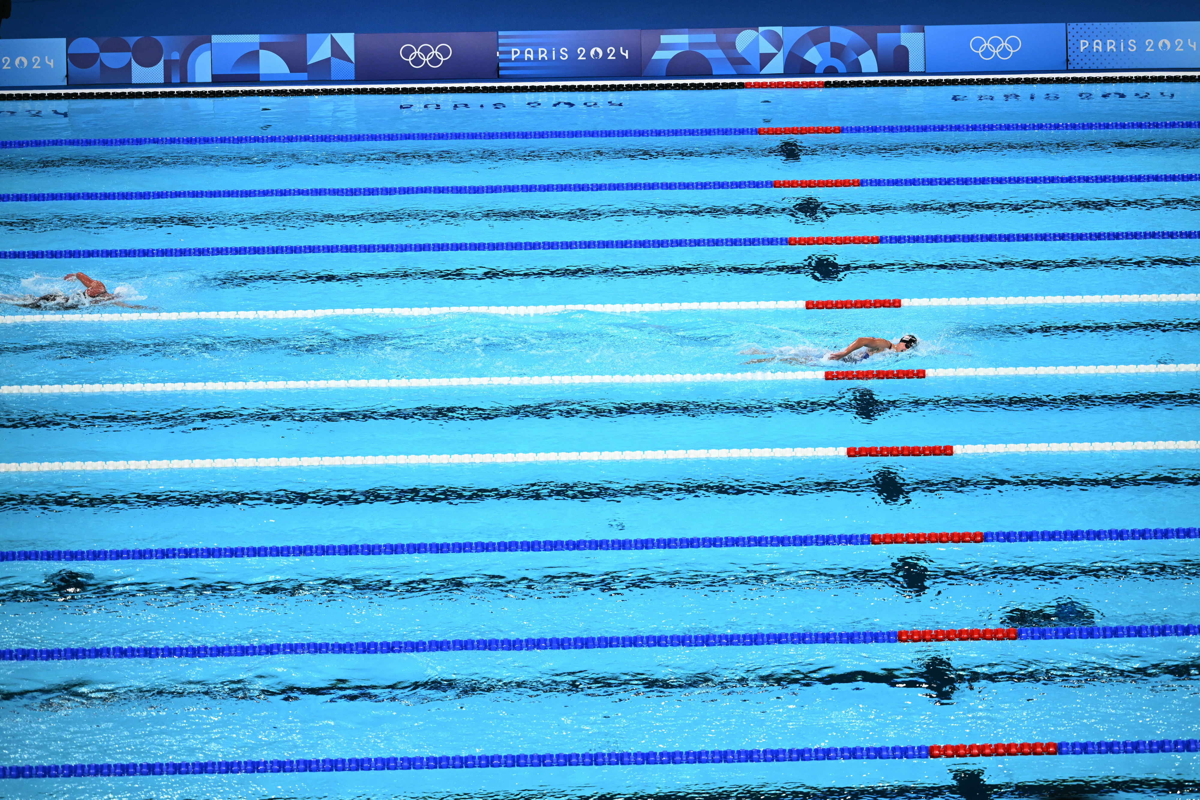 Three competitive swimmers in a race in an Olympic swimming pool, with Paris 2024 signage visible
