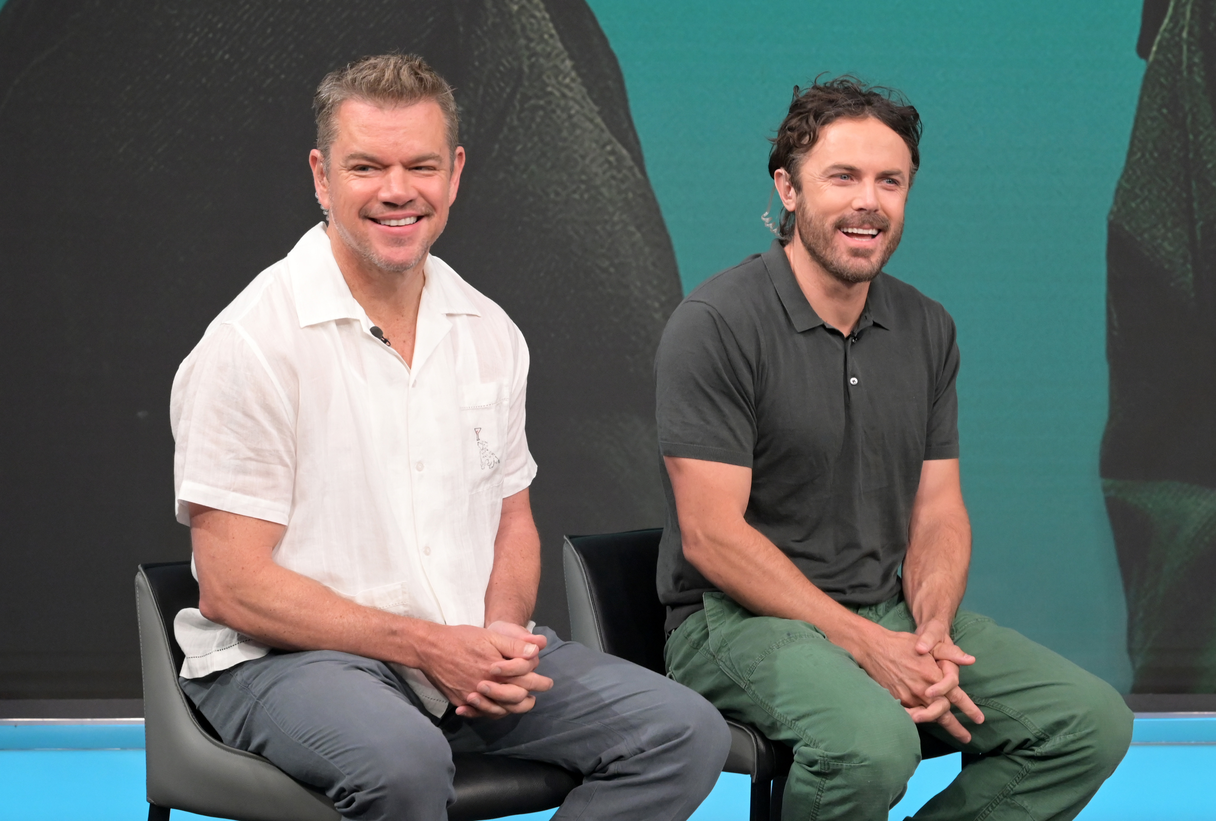 Matt Damon and Casey Affleck sit on stage, smiling and engaged in conversation. Matt wears a white shirt, and Casey wears a dark polo with green pants