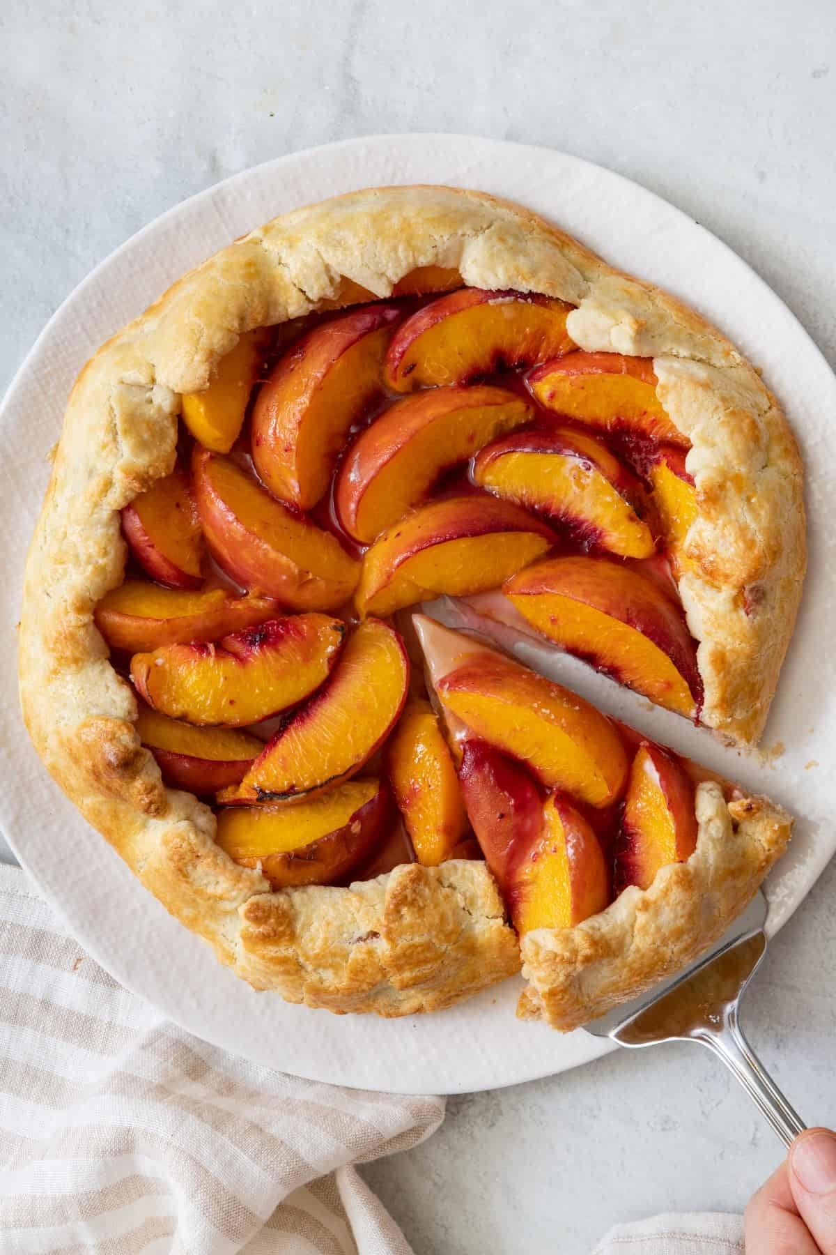 A peach galette on a white plate with a slice being lifted by a server. The rustic crust surrounds sweetly arranged peach slices