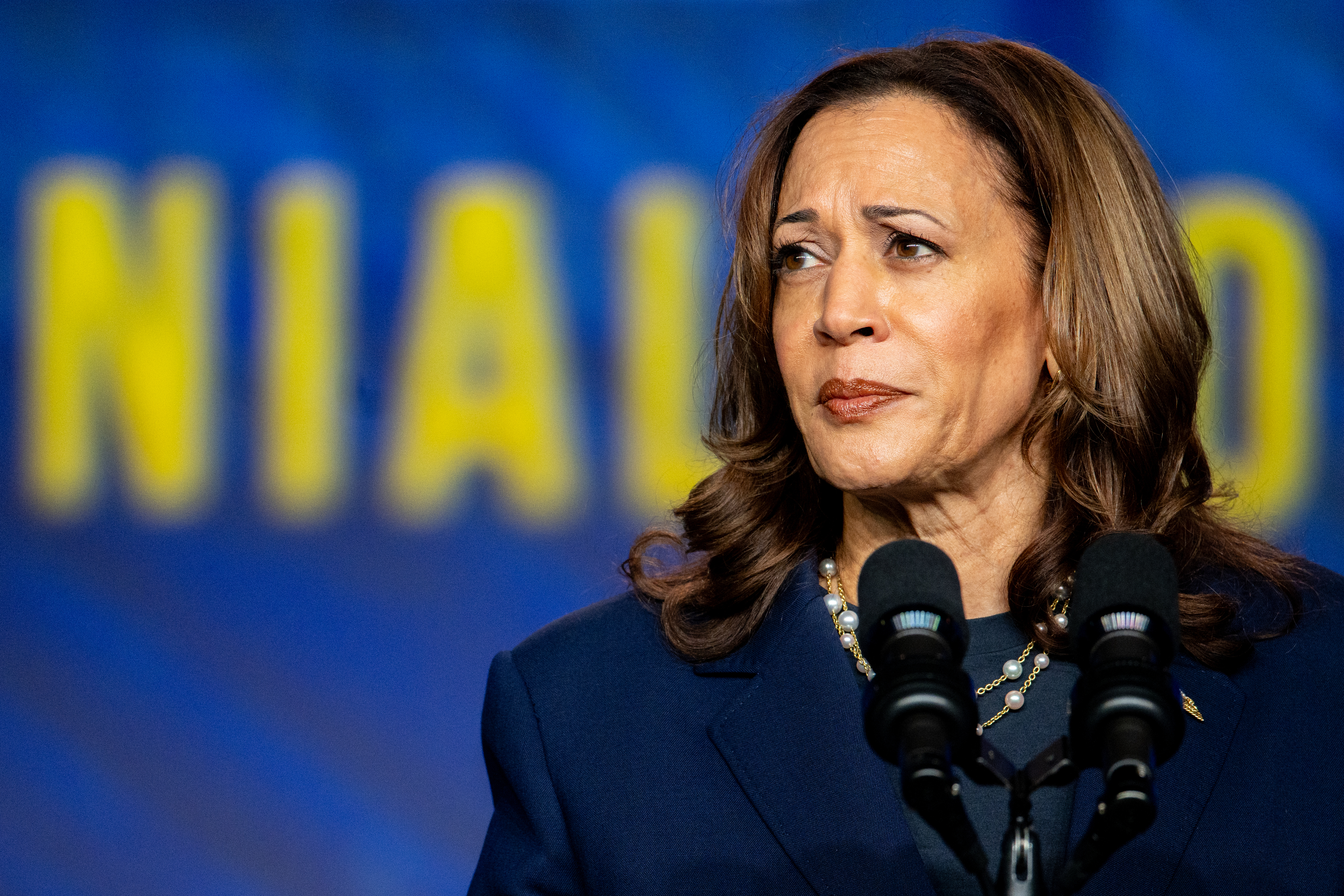 Kamala Harris, wearing a dark blazer and pearl necklace, speaks at a podium with a blue and yellow backdrop
