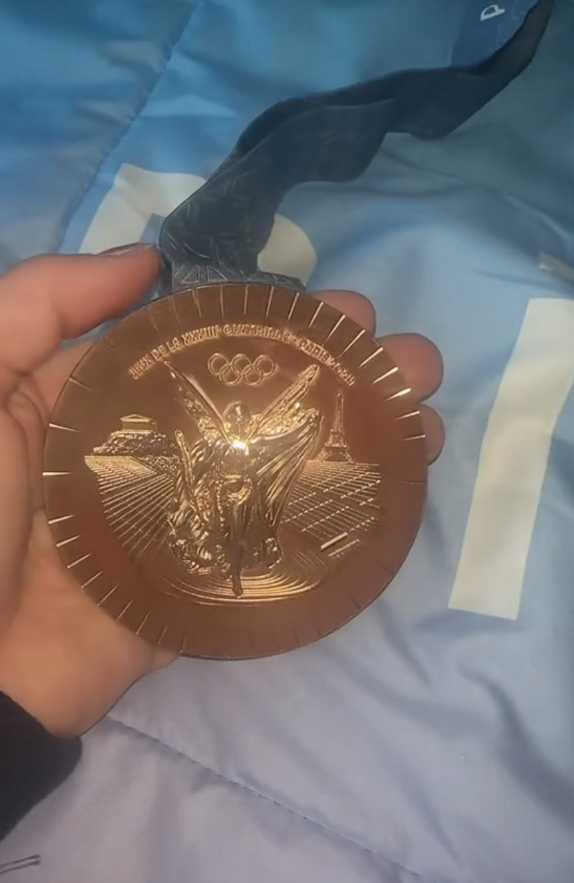 Hand holding an Olympic bronze medal with the Olympic rings and a seated winged figure on the front