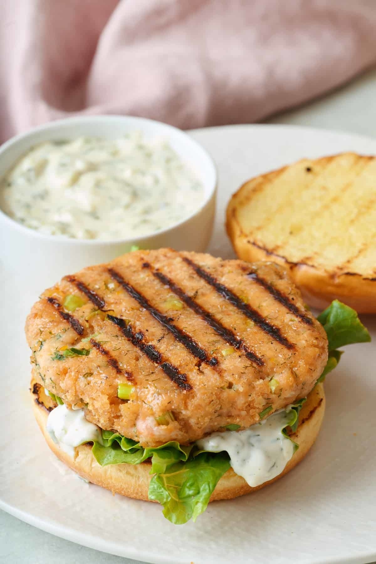 Salmon burger with grilled patty, lettuce, and sauce on a toasted bun. A bowl of tartar sauce is on the side