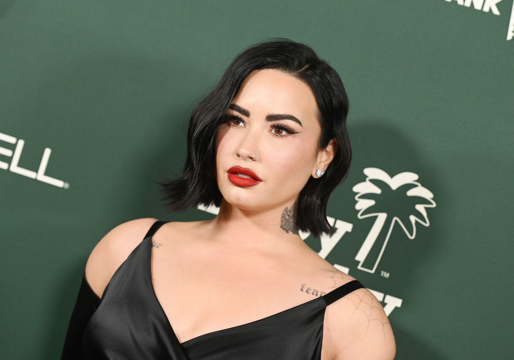Demi Lovato poses at an event, wearing a sleek, black satin dress with a V neckline. She has short, wavy hair and wears bold red lipstick