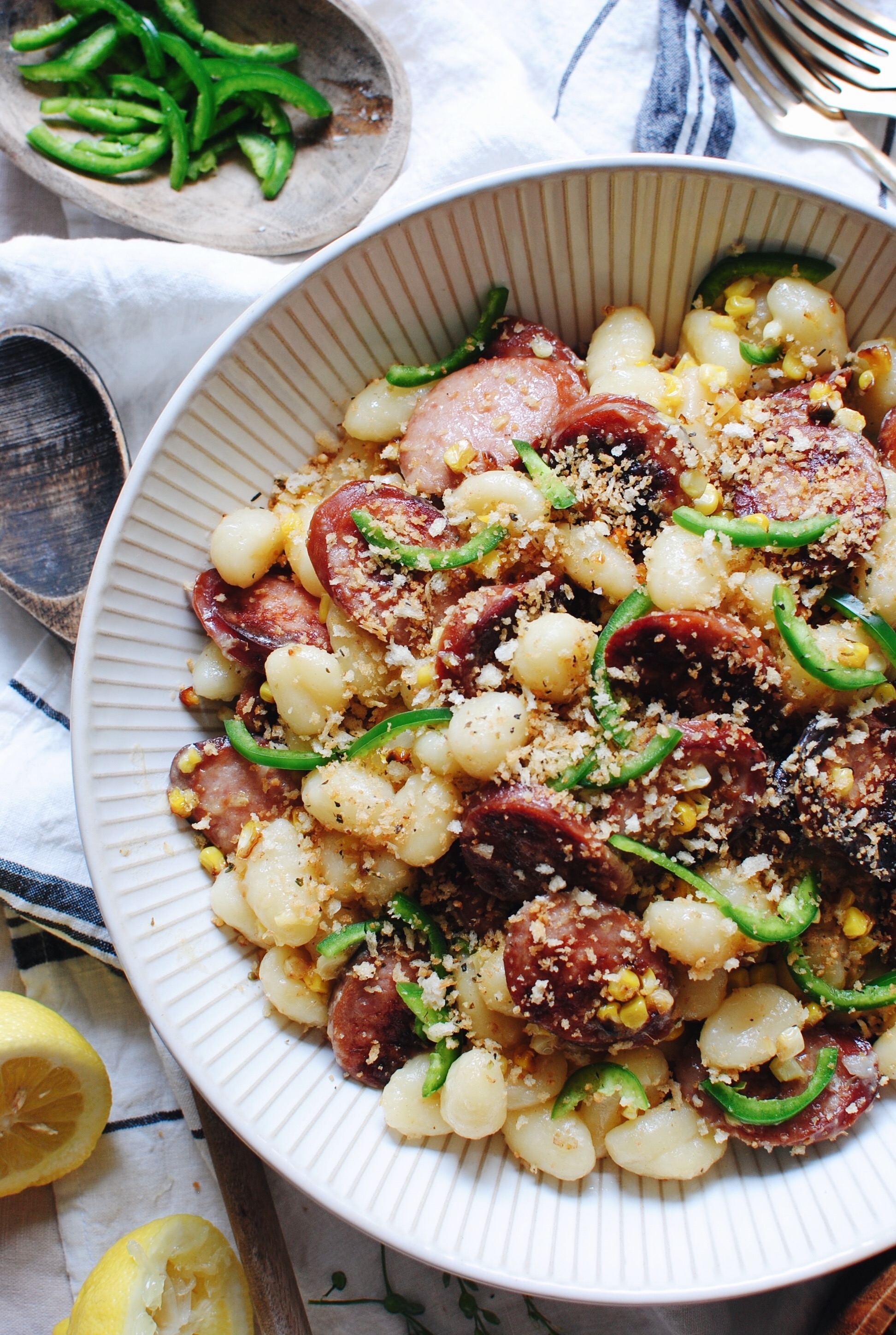 A bowl of gnocchi mixed with sliced sausage, corn, jalapeño slices, and topped with breadcrumbs. Sliced jalapeños and lemon wedges are on the side