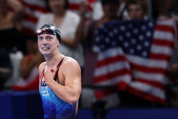 Katie Ledecky in a swim cap and swimsuit, smiling and holding her shoulder with USA flag in the background at a swimming event