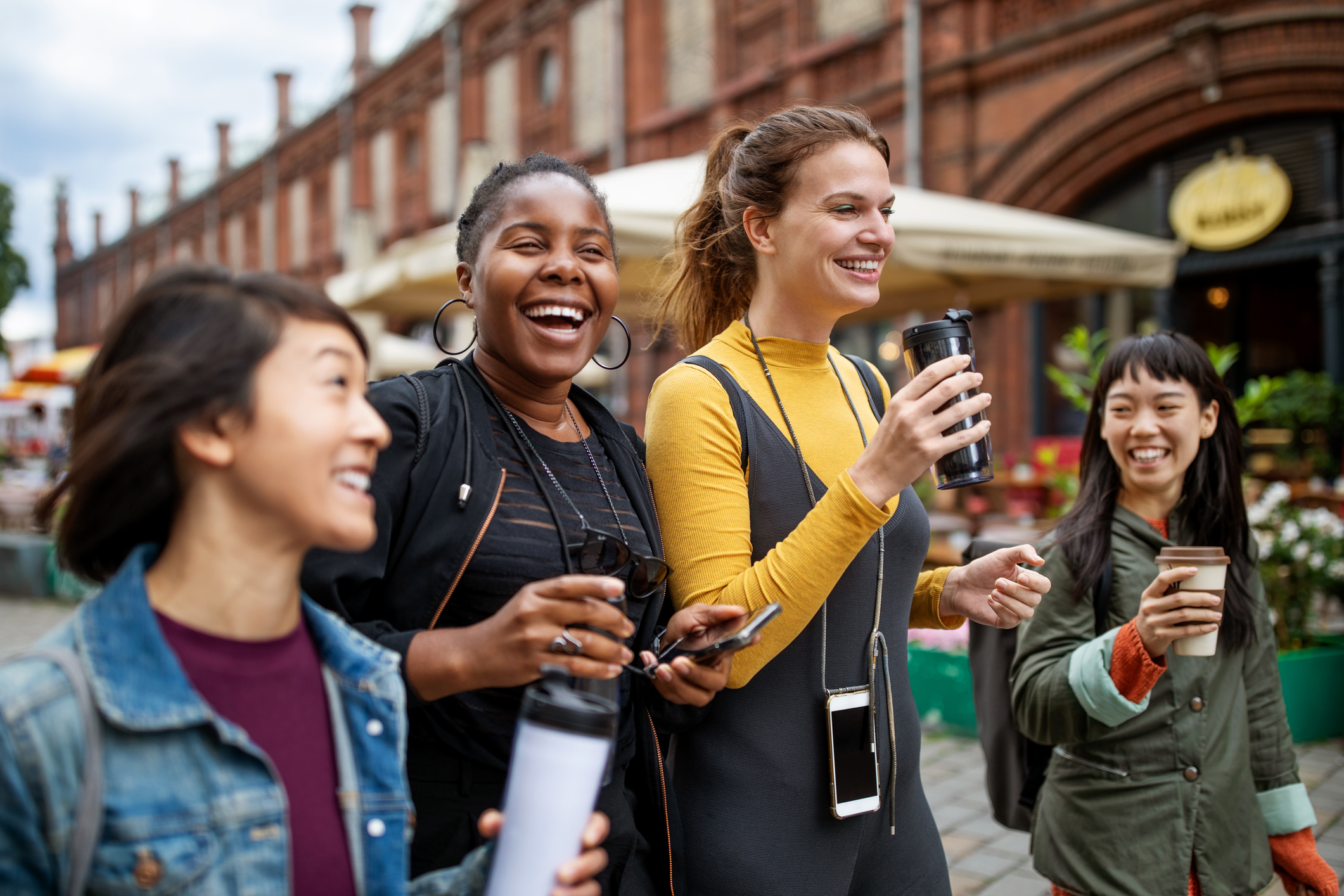 A group of four smiling people walk outside holding coffee cups, suggesting a joyful travel experience. Brick building in the background