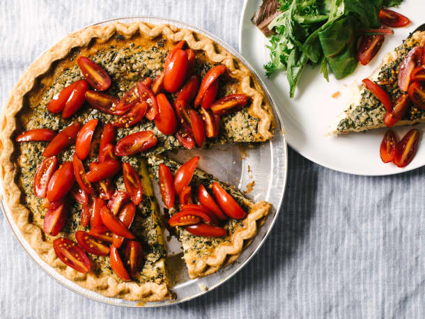 A freshly baked vegetable quiche topped with sliced cherry tomatoes next to a plate with a slice of the quiche and some mixed greens
