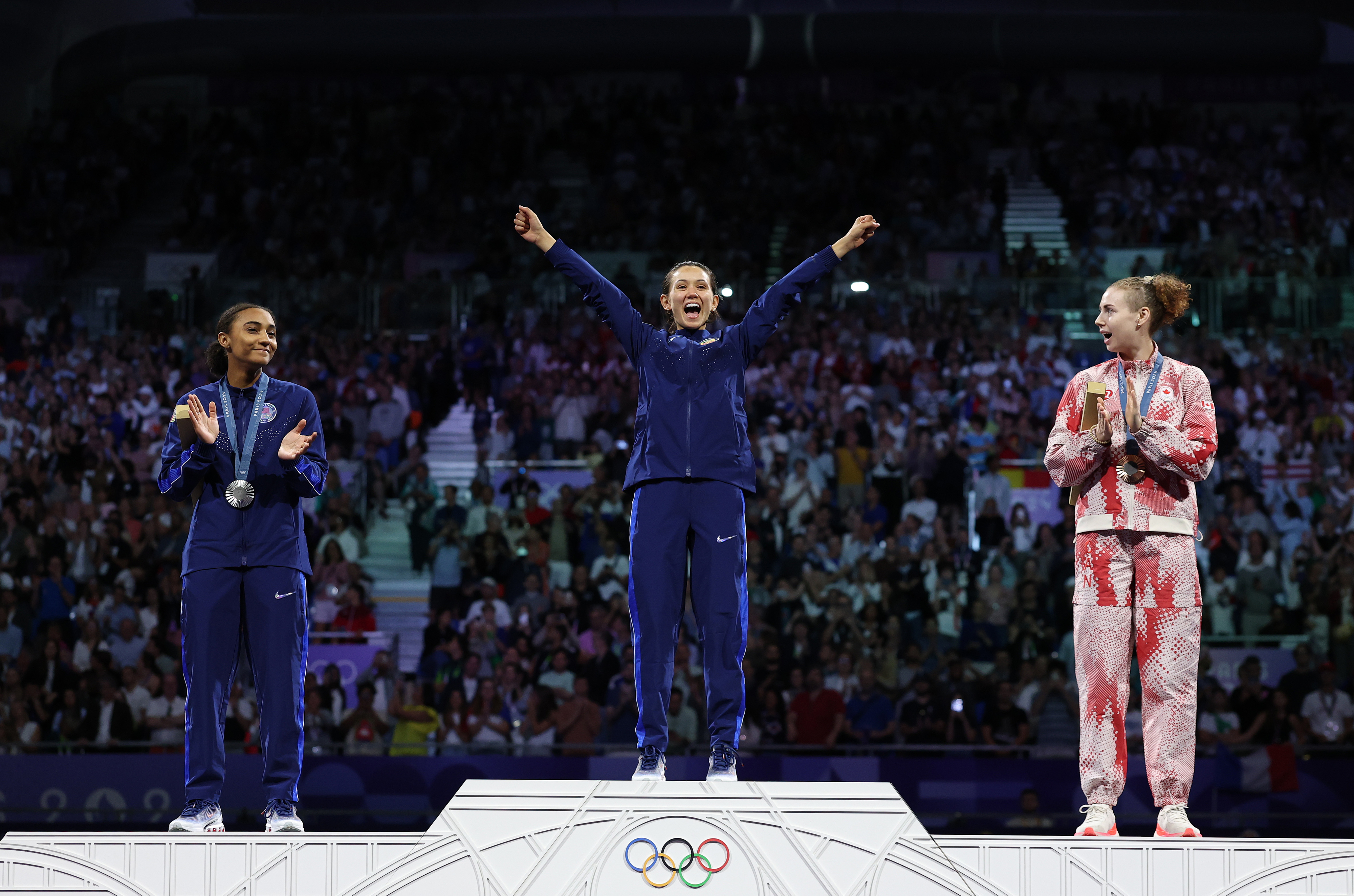 Athletes celebrate on the Olympic podium: the gold medalist in the center is smiling with arms raised, while the silver and bronze medalists clap