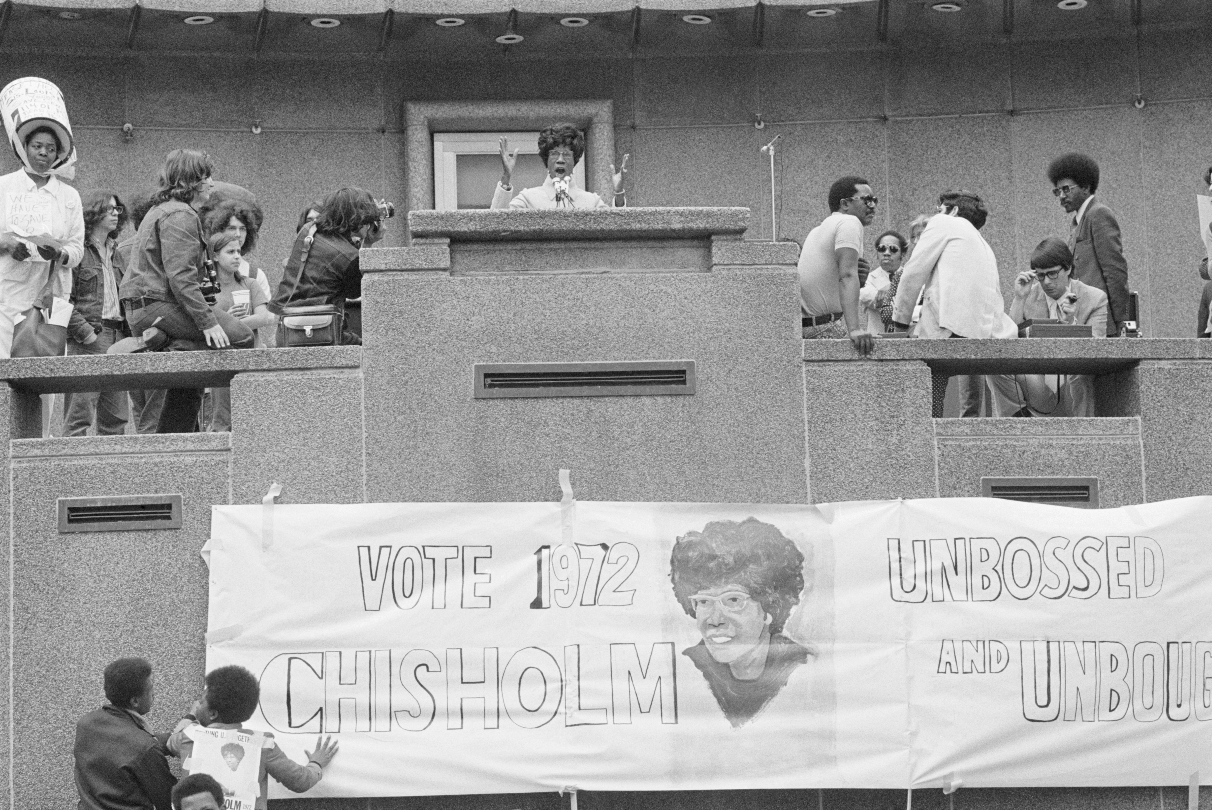 Shirley Chisholm giving a speech on an elevated stage with a "Vote 1972 Chisholm Unbossed and Unbought" banner featuring her portrait below her
