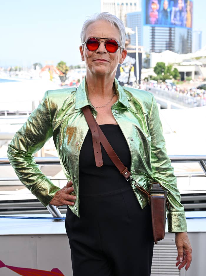 Jamie Lee Curtis poses outdoors wearing a shiny jacket, sunglasses, and a crossbody bag