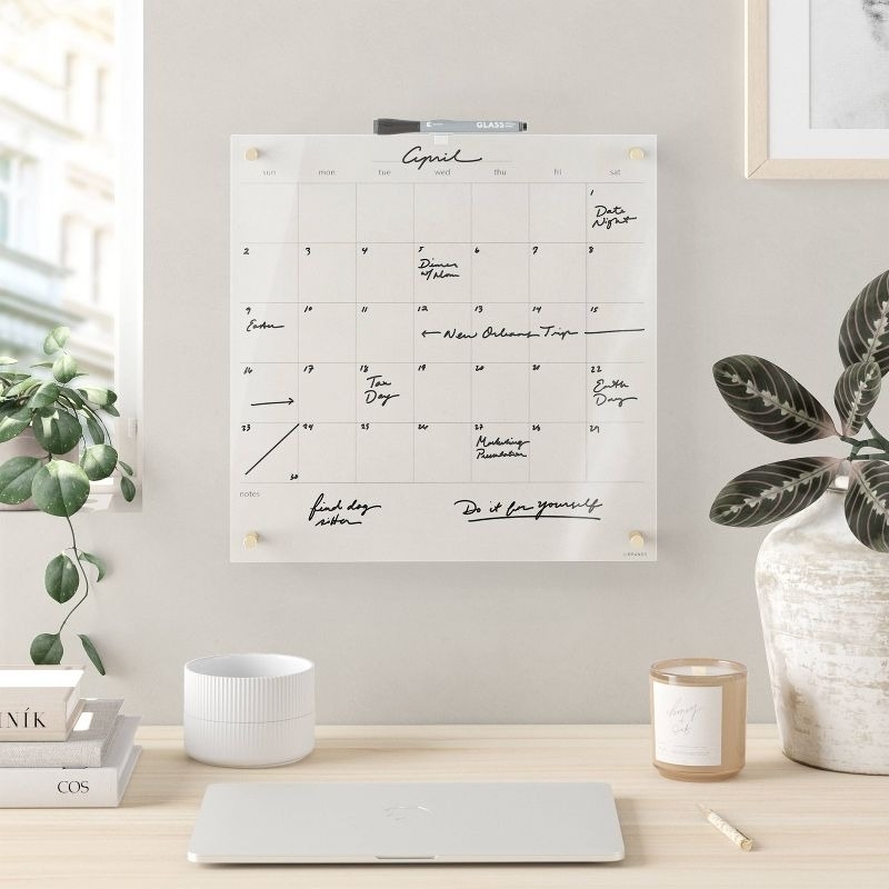 Monthly calendar on wall with handwritten notes, desk below with laptop, candle, books, and a potted plant. Scene in an organized and minimalistic workspace