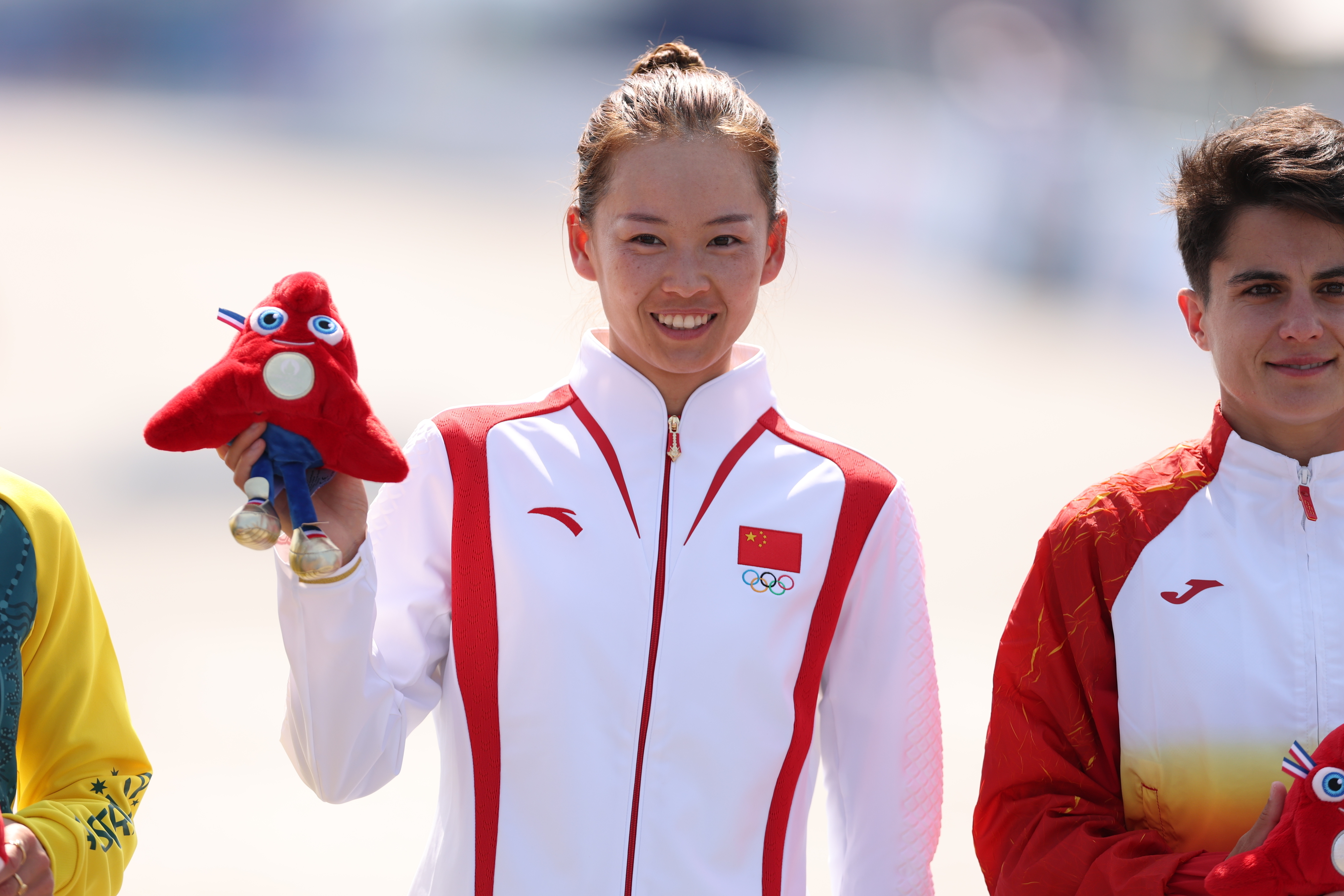 Athlete in a red and white tracksuit holding a plush toy alongside two other athletes in tracksuits at a sports event