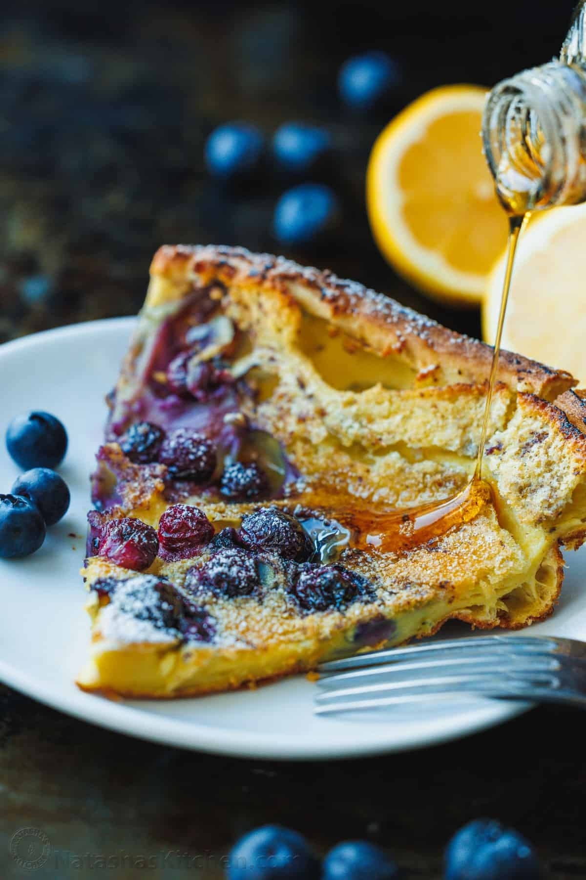 A plated slice of blueberry pancake pie being drizzled with syrup, accompanied by fresh blueberries and a lemon half