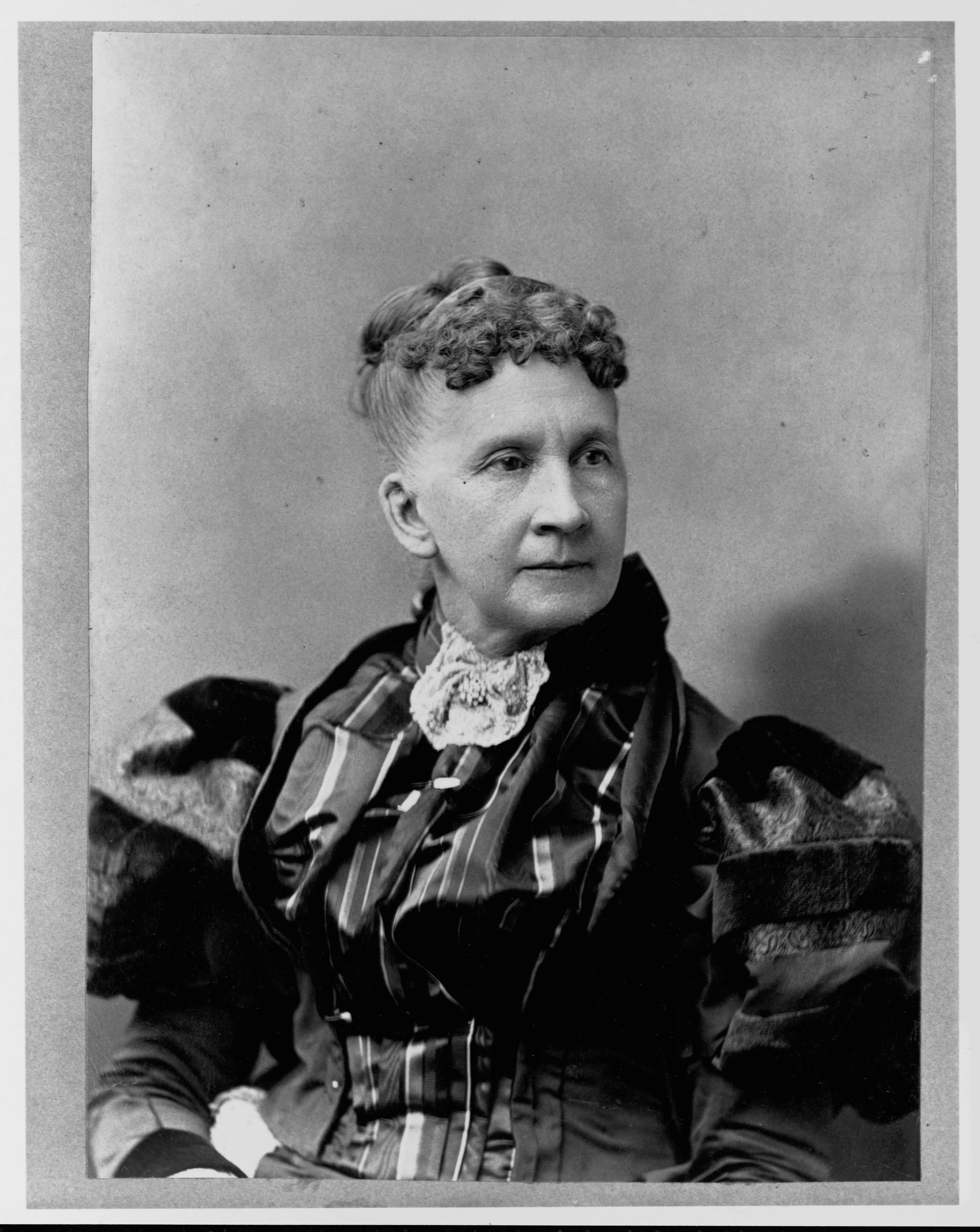 A woman with curly hair styled up, wearing a high-collared blouse and a dress with puffed sleeves, looks off-camera