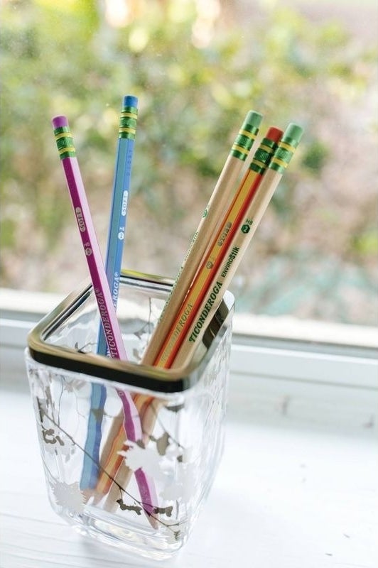 A glass container with colorful pencils is placed on a windowsill in a bright, natural light setting
