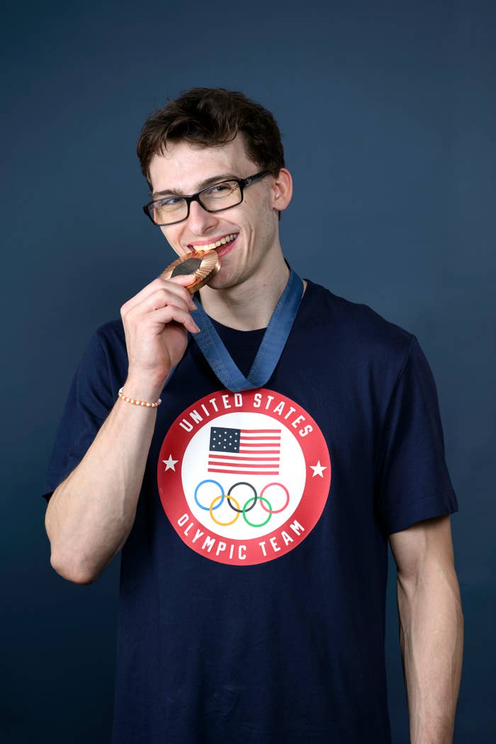 A man wearing a "United States Olympic Team" shirt smiles while holding a medal in his mouth