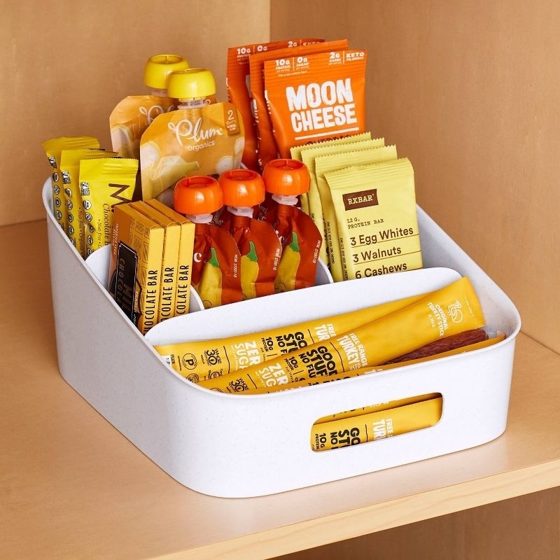 Assorted healthy snacks, including Plum Organics pouches, Moon Cheese packets, and RXBARs, neatly arranged in a white container on a wooden shelf