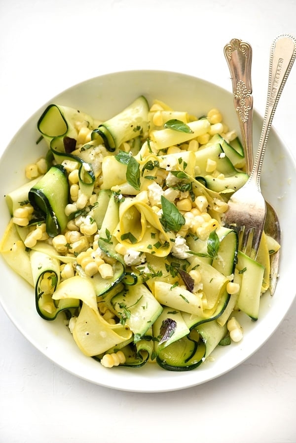 A bowl of zucchini and corn salad garnished with fresh herbs, with a fork and spoon placed inside the dish