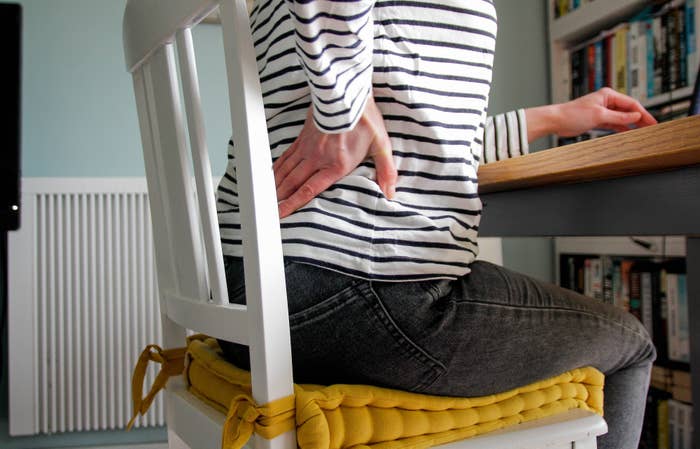 A person sitting on a cushioned chair, wearing a striped shirt and jeans, is holding their lower back, possibly indicating discomfort or pain