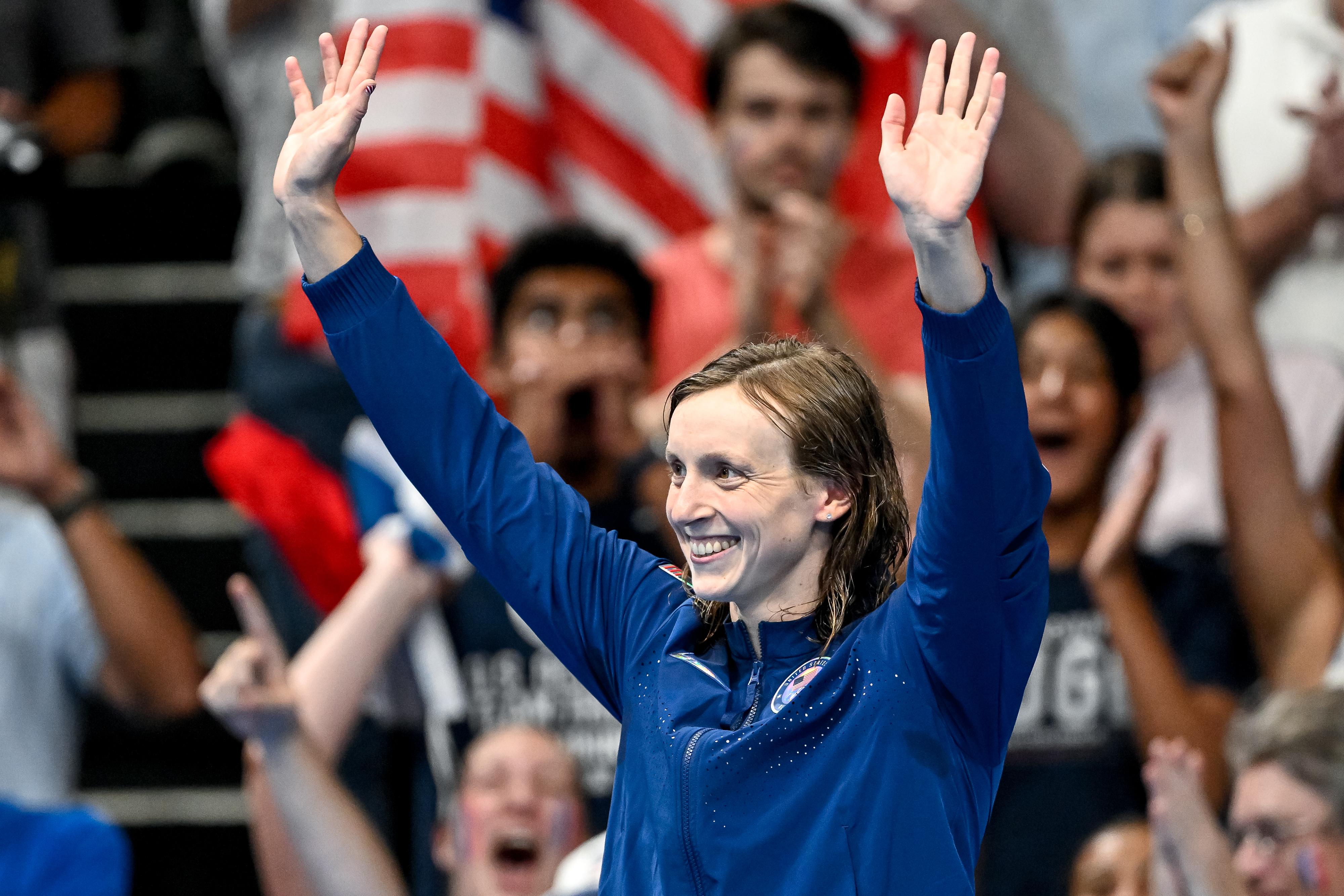 Katie Ledecky smiles with arms raised in celebration, wearing a sports jacket. A crowd of cheering fans is in the background holding flags