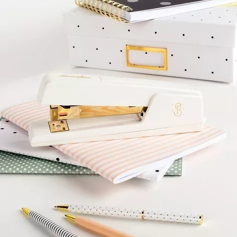 White stapler on a stack of patterned notebooks and stationary, with a white and gold polka-dotted box and gold-rimmed pen and pencil in the foreground