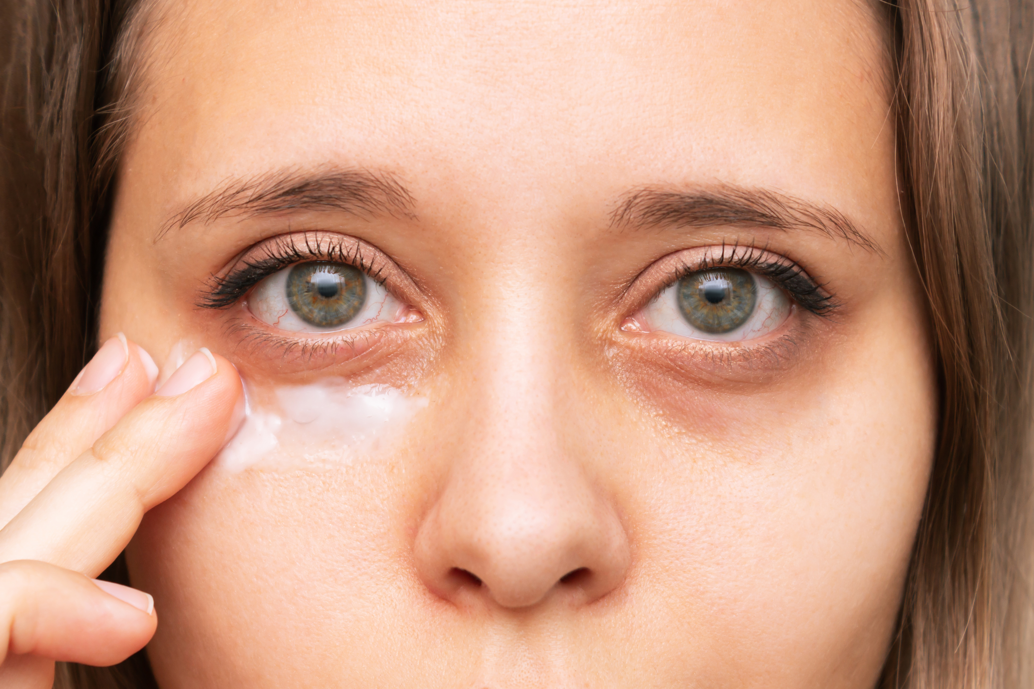 A close-up of a woman applying cream under her eye. The woman's face is prominently shown with visible under-eye circles
