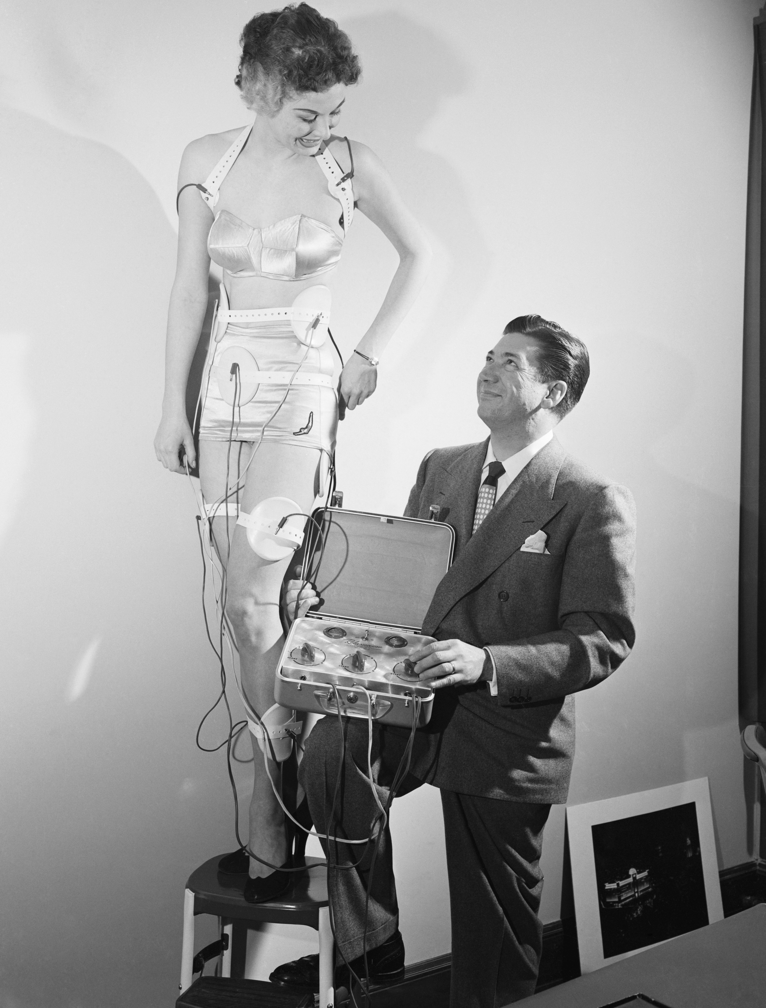 A woman stands on a stool with wires connected to it. A man in a suit holds a suitcase-like device that the wires lead to.