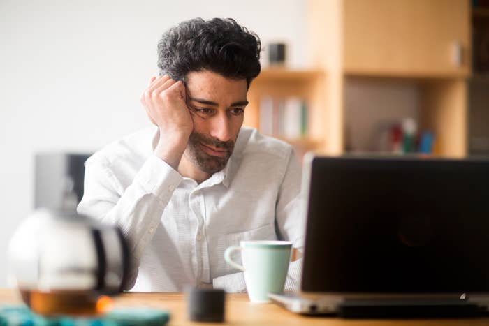 Man in a white shirt looking at a laptop, resting his head on his hand, with a coffee mug and blurred background items nearby