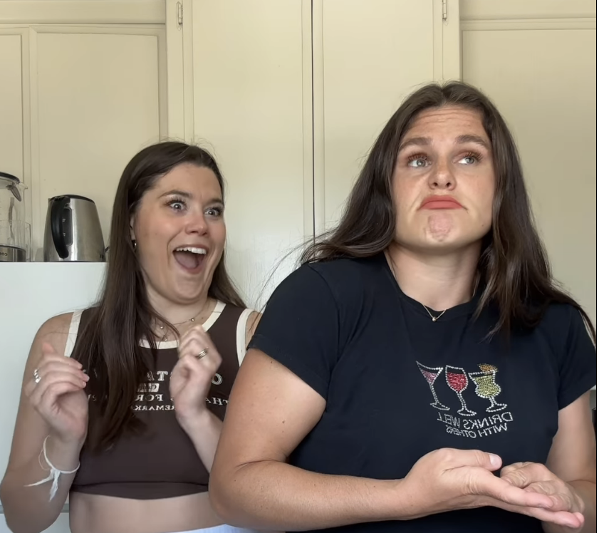 Olivia and Ilona stand in a kitchen; Olivia on the left is smiling excitedly while Ilona on the right looks puzzled