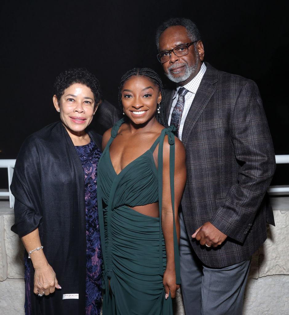 Simone Biles stands between her parents, Nellie and Ron, all smiling. Simone wears an elegant gown, while her mother and father are dressed in formal attire