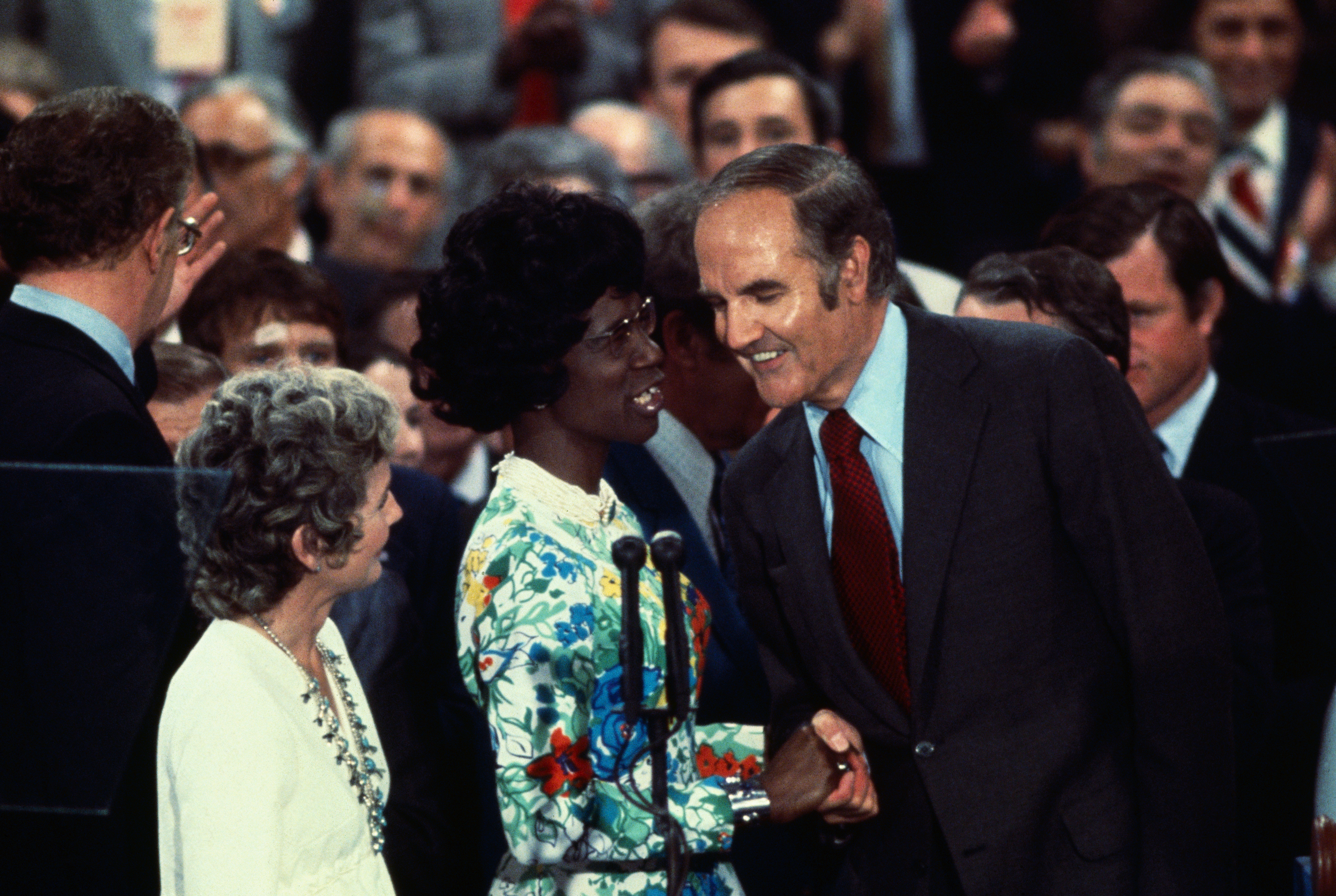 Shirley Chisholm wearing a floral-patterned dress shakes hands with George McGovern, while a crowd watches. Another person stands beside them
