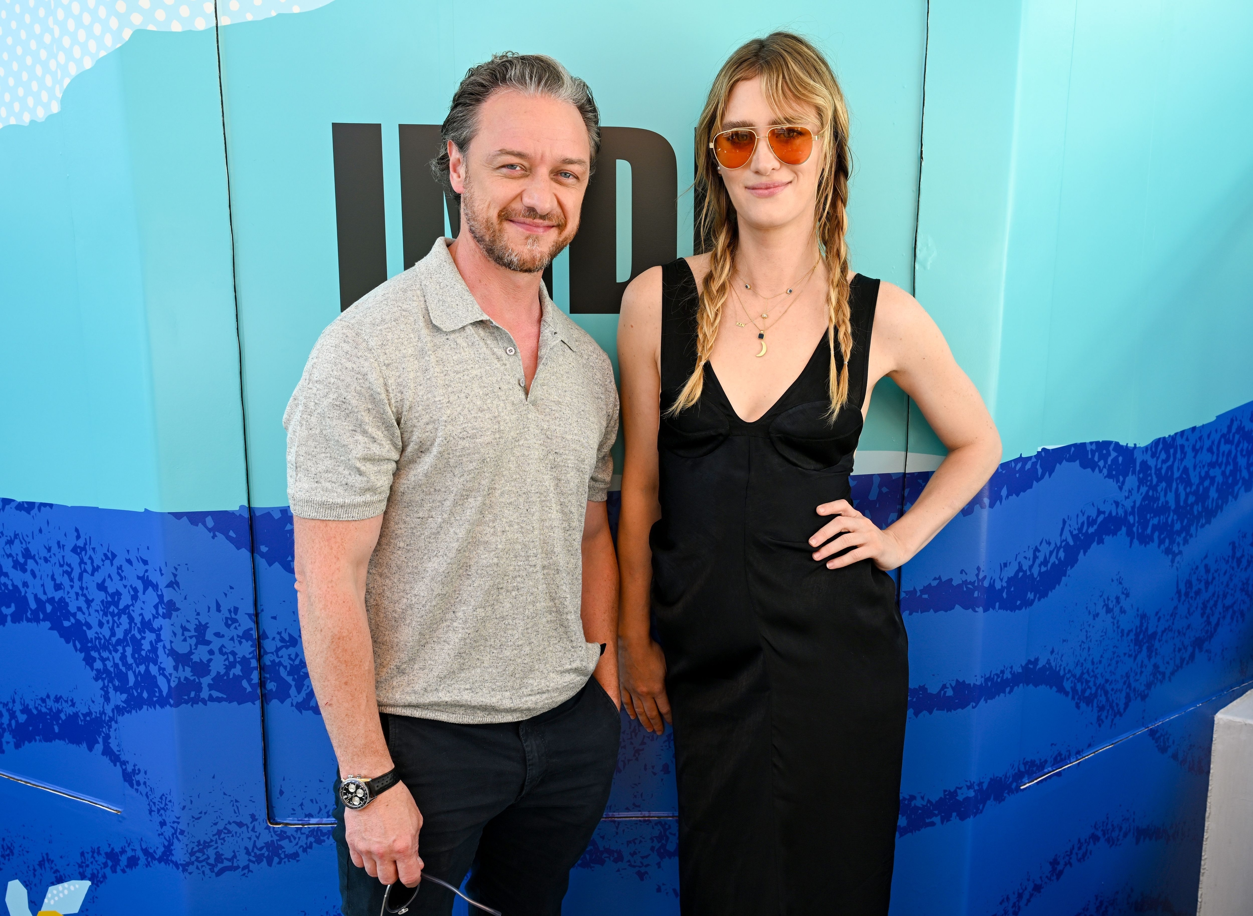 James McAvoy and Mackenzie Davis pose together at an event. McAvoy wears a casual shirt and jeans, while Debicki dons a black dress with braids and sunglasses