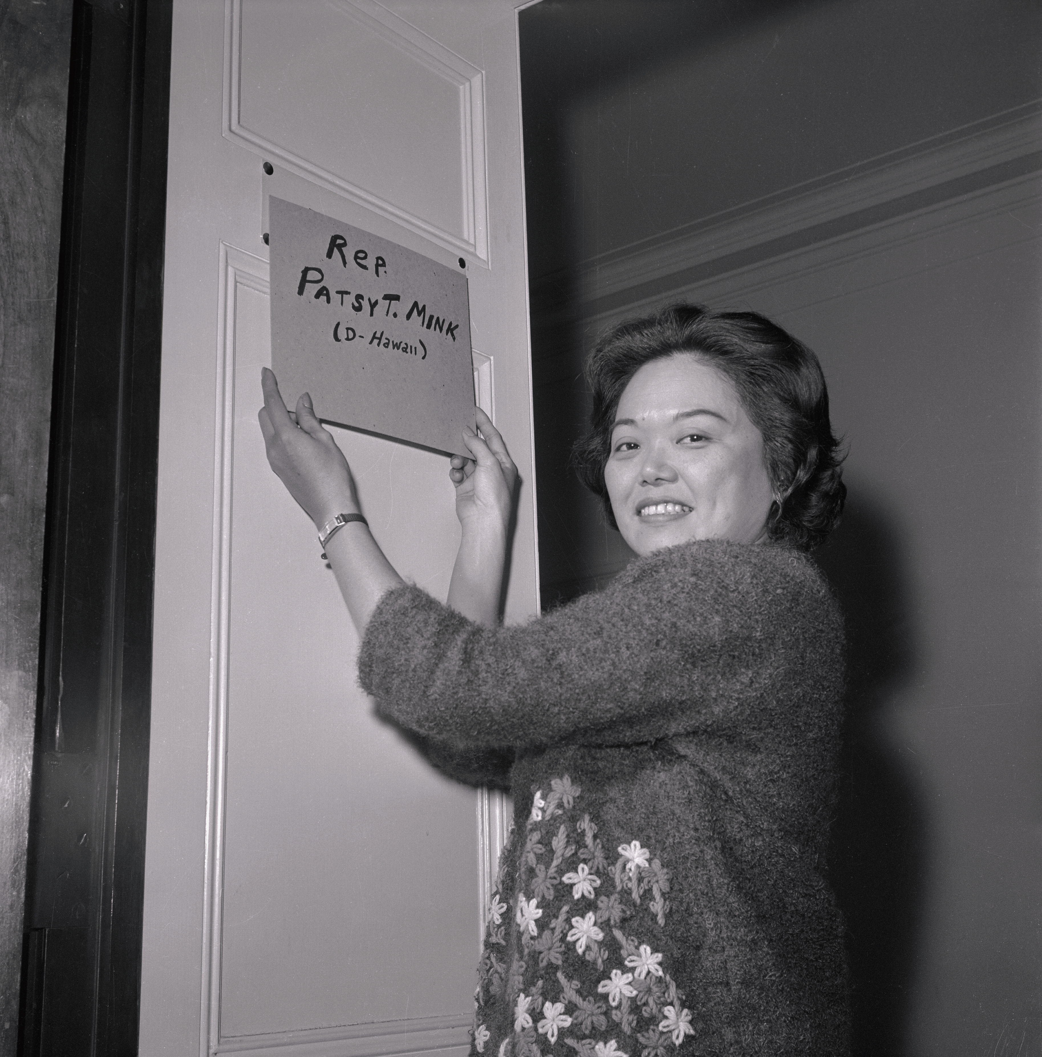 Patsy T. Mink, with a cheerful expression, is seen hanging a sign that reads "Rep Patsy T. Mink (D-Hawaii)" on a door