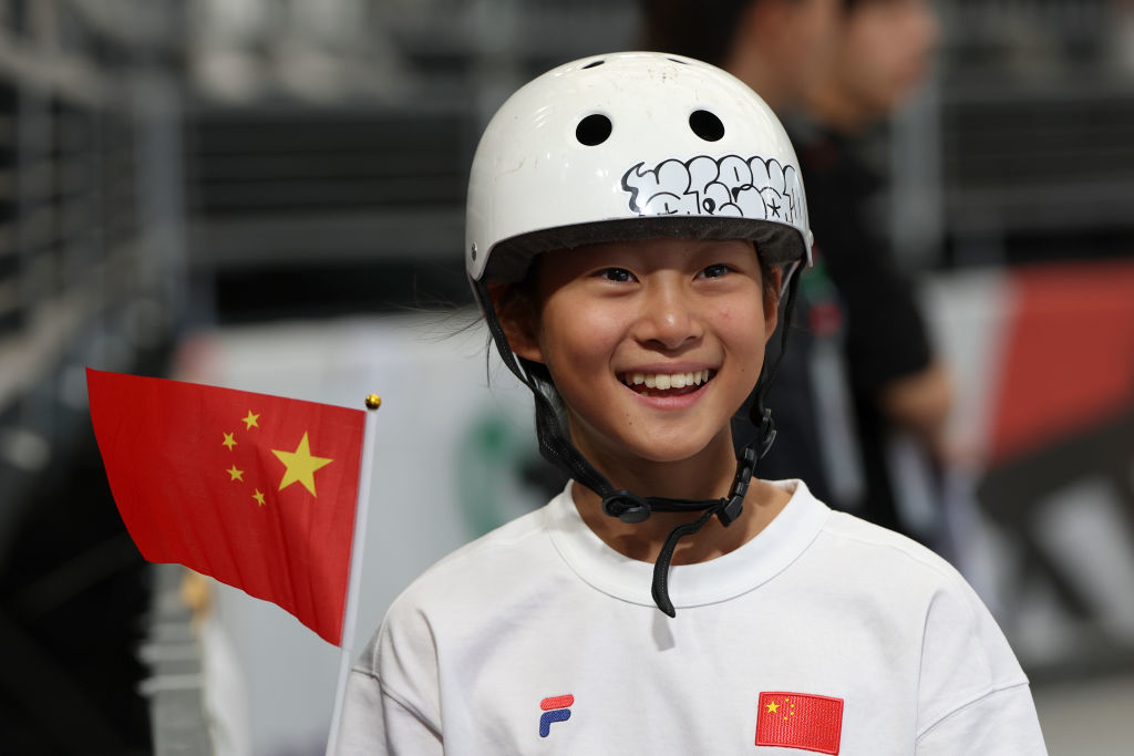 A young girl, smiling while wearing a skateboard helmet adorned with stickers, holds a small Chinese flag. She is dressed in a white athletic top