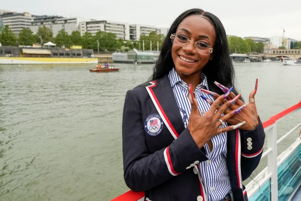 Sha'Carri Richardson smiles on a boat, wearing a striped shirt, navy blazer with USA Olympics patch, and long, decorated nails