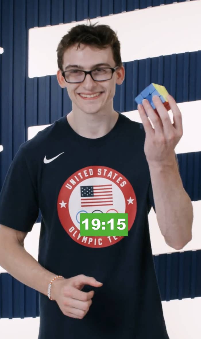 Stephen wearing a United States Olympic Team shirt smiles while holding a solved Rubik's Cube. A timer on the image shows 19:15