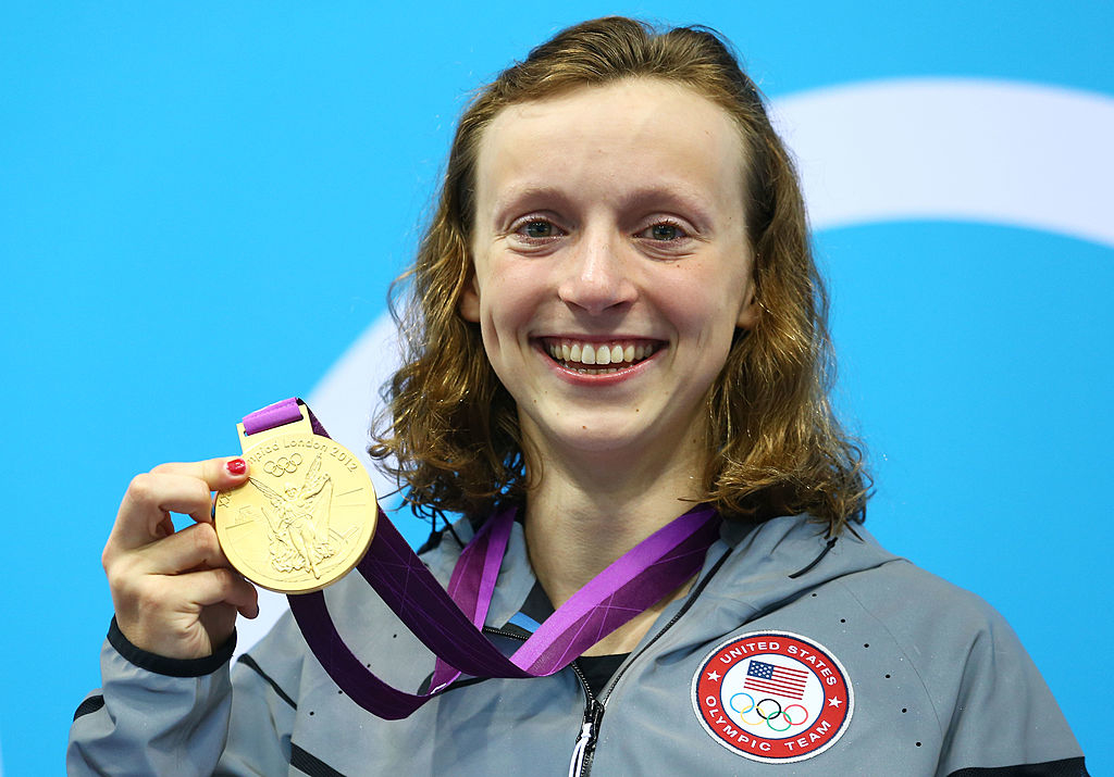 Katie Ledecky smiles and holds up her Olympic gold medal while wearing a Team USA jacket