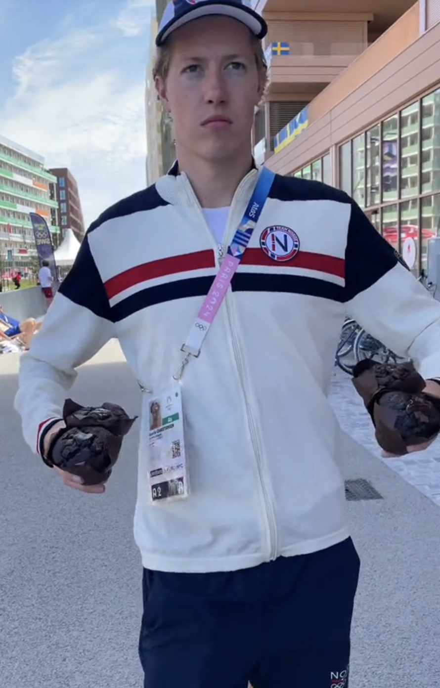 Henrik wearing a jacket with stripes, a cap, and holding four large chocolate muffins near his waist in Olympic Village