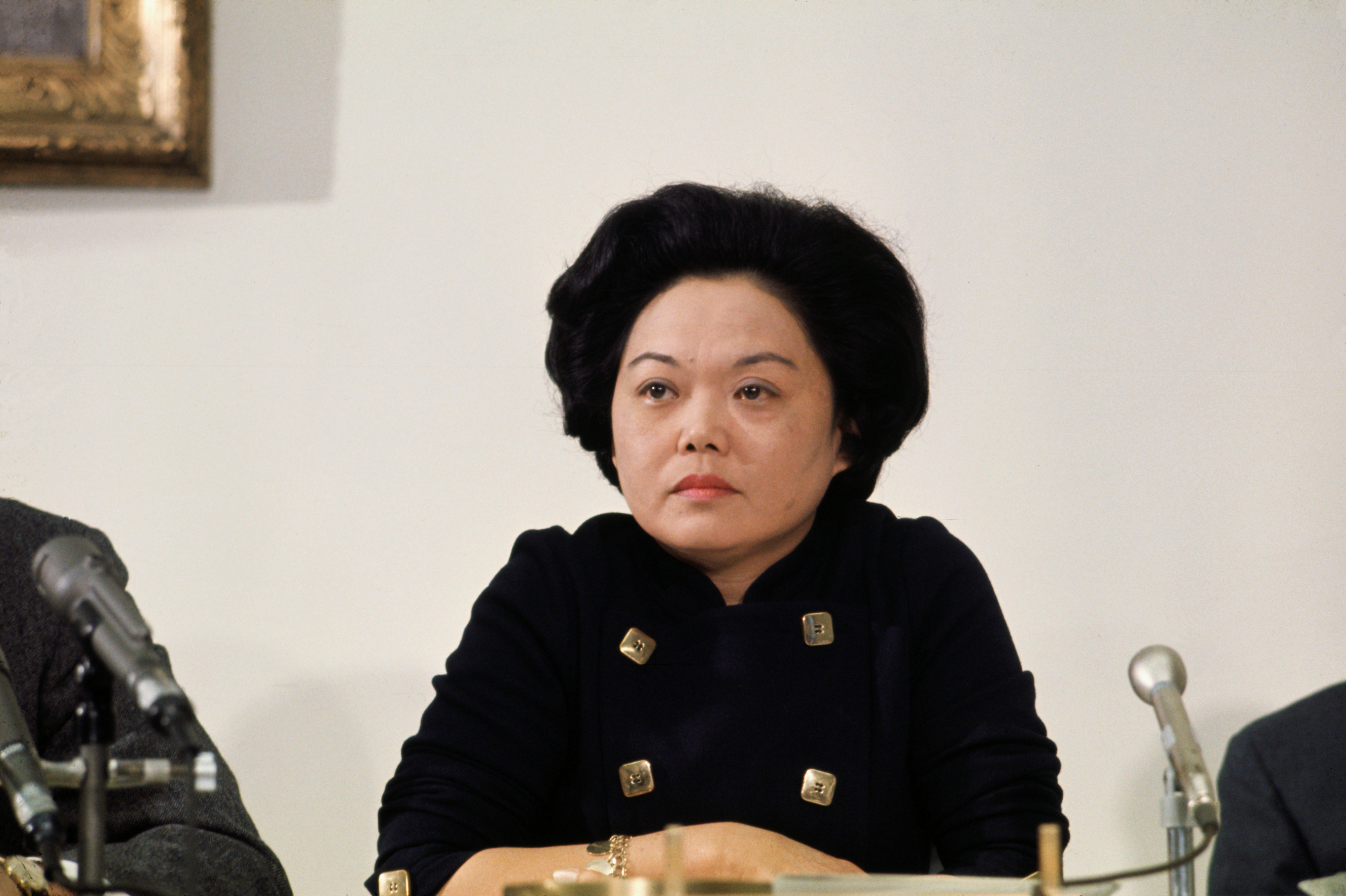 Woman with shoulder-length black hair, wearing a double-breasted dress, sits at a table with microphones