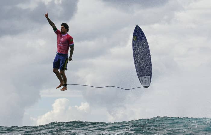 Gabriel Medina is surfing, caught mid-air with one hand pointing up and his surfboard vertically above the water