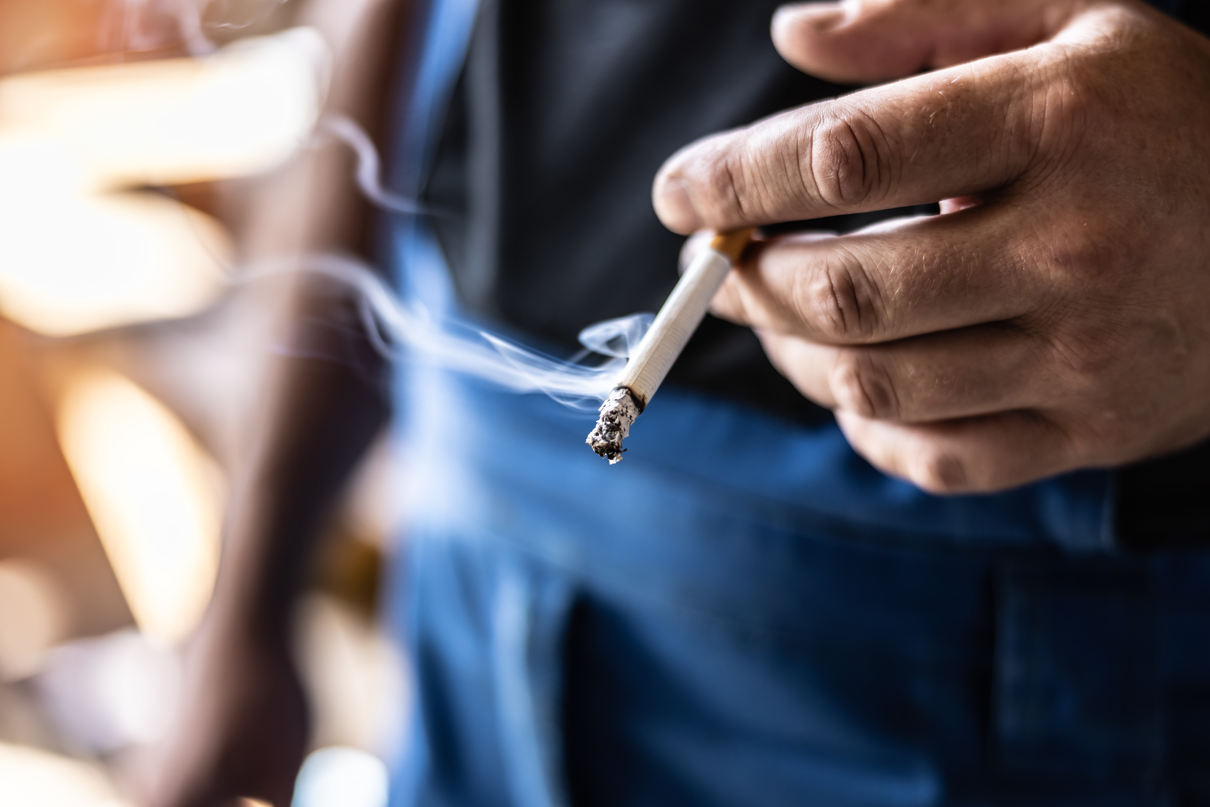 A hand holding a lit cigarette with smoke rising from the cigarette's tip. The person is wearing blue pants