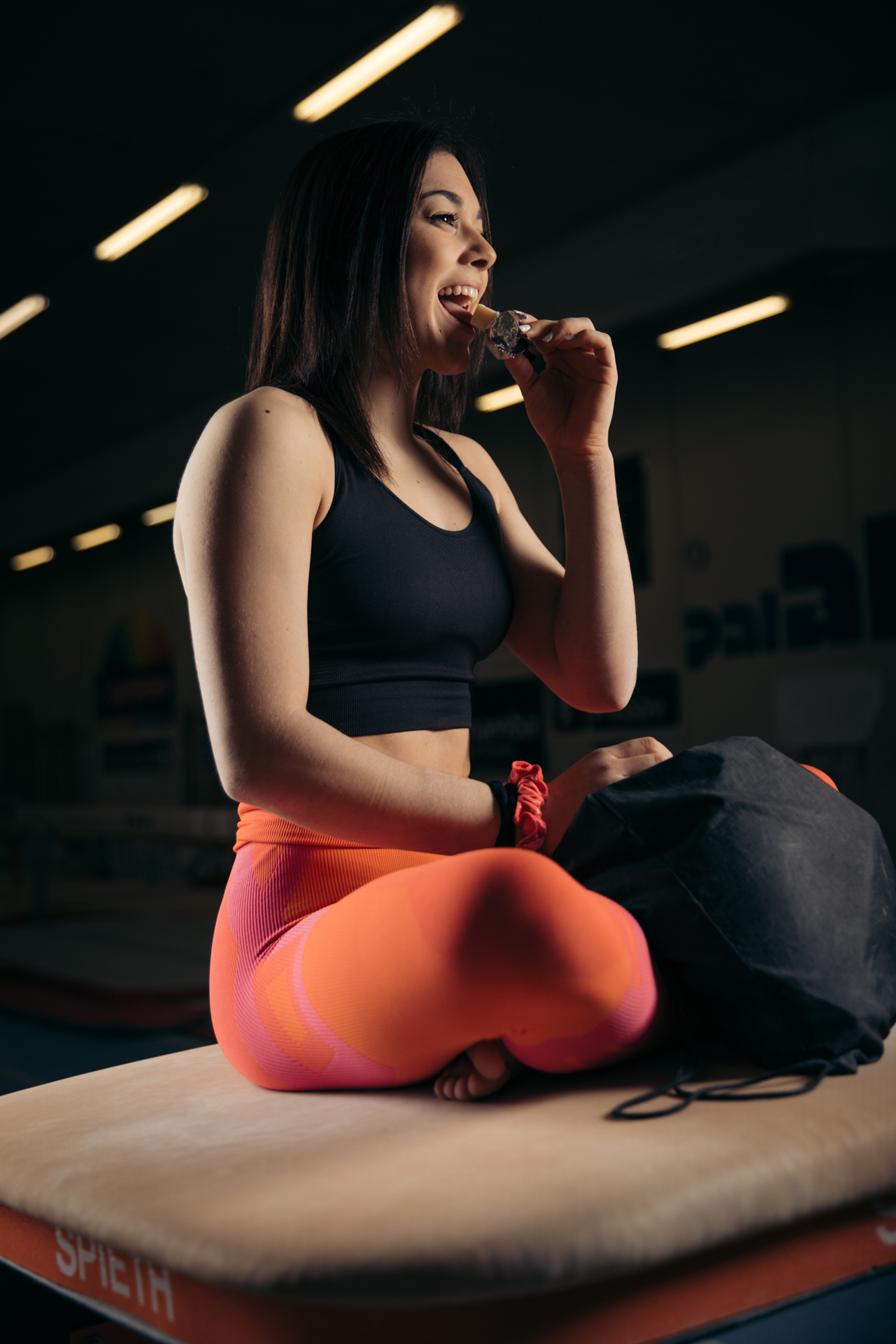 A person is sitting cross-legged on a bench, wearing a black sports bra and bright workout pants, smiling while eating a snack bar, with a gym in the background