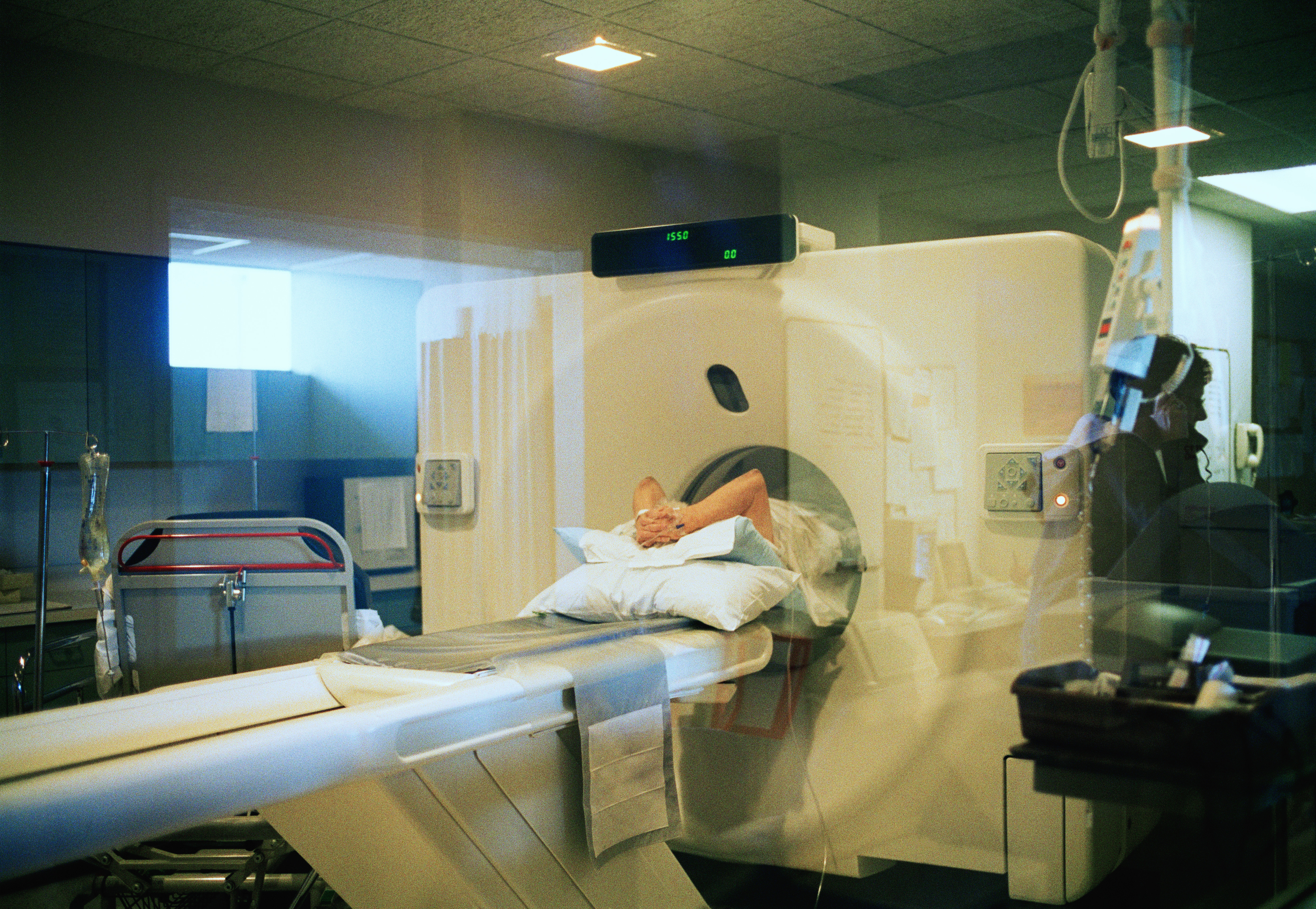A person is lying down on a medical scanner bed, in a hospital room, preparing for an MRI or CT scan. Hospital equipment is visible around them