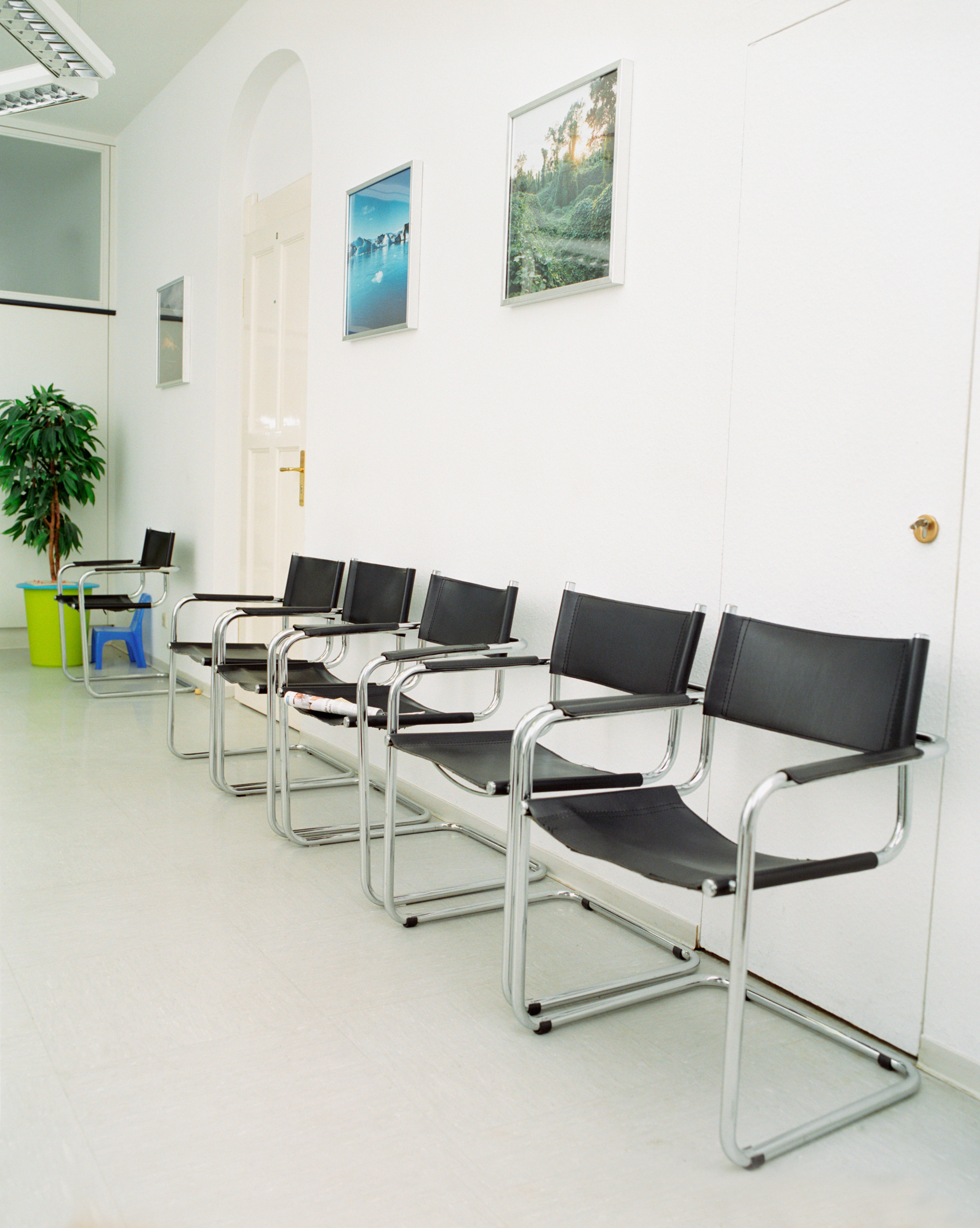 Row of empty black chairs with metal frames in a waiting area, white walls with framed photos, small plant, and a closed door in the background