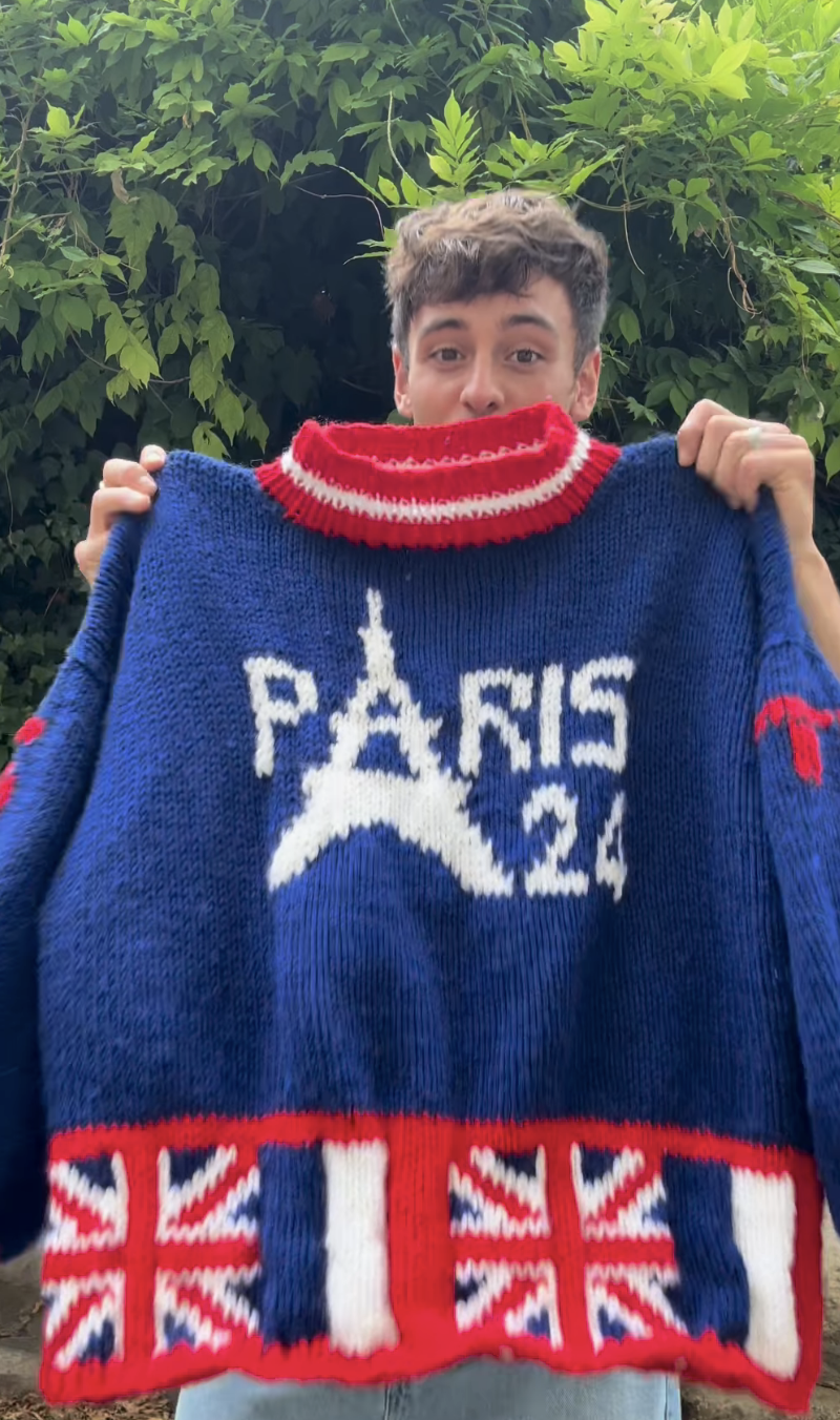 Tom Daley holds a knitted sweater with "PARIS 24" and a tower design, indicating the 2024 Paris Olympics. The sweater also features flag motifs at the bottom